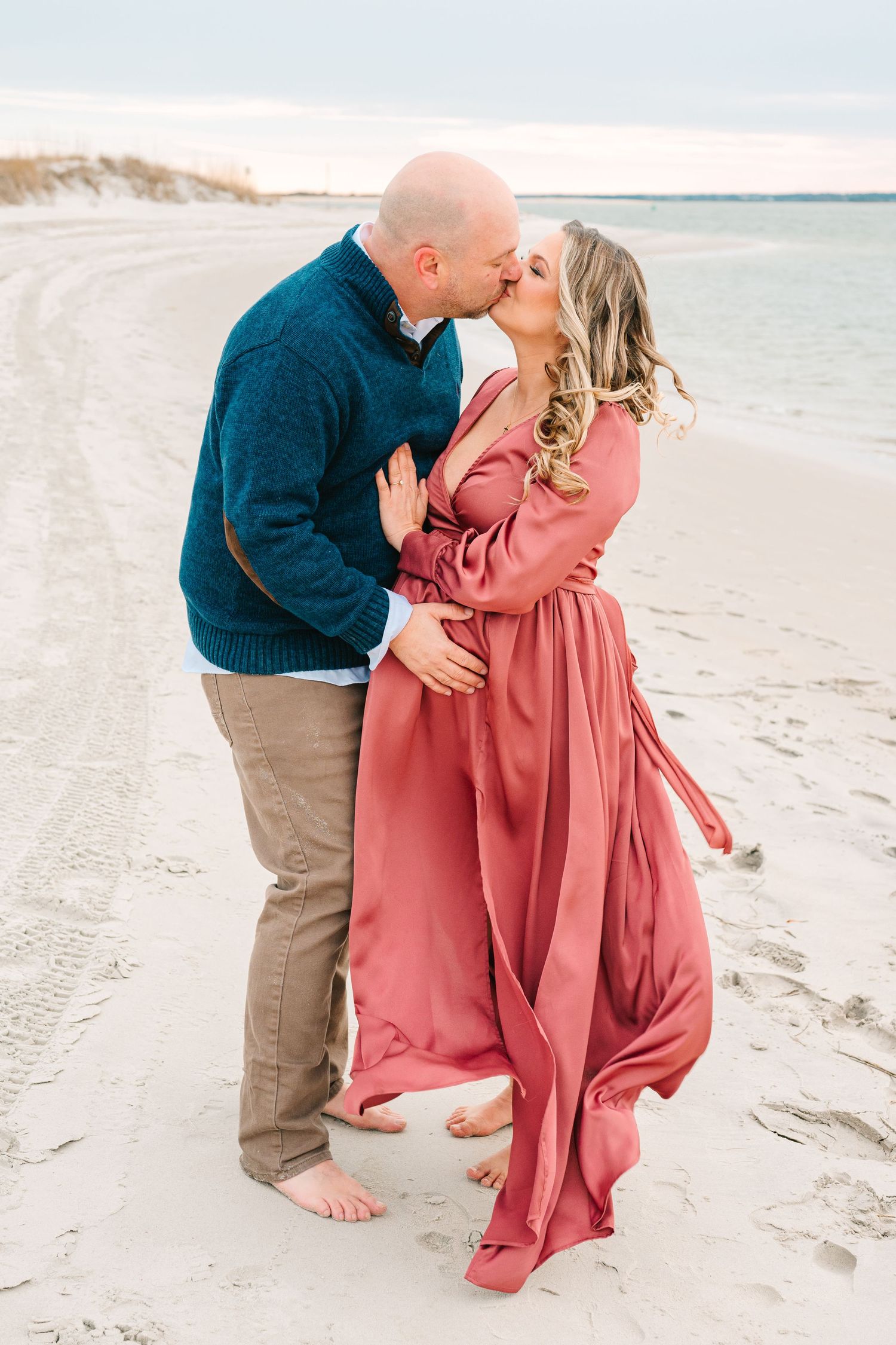 A romantic couple embraces and kisses on a sandy beach, with the woman wearing a flowing pink dress and man in navy sweater.