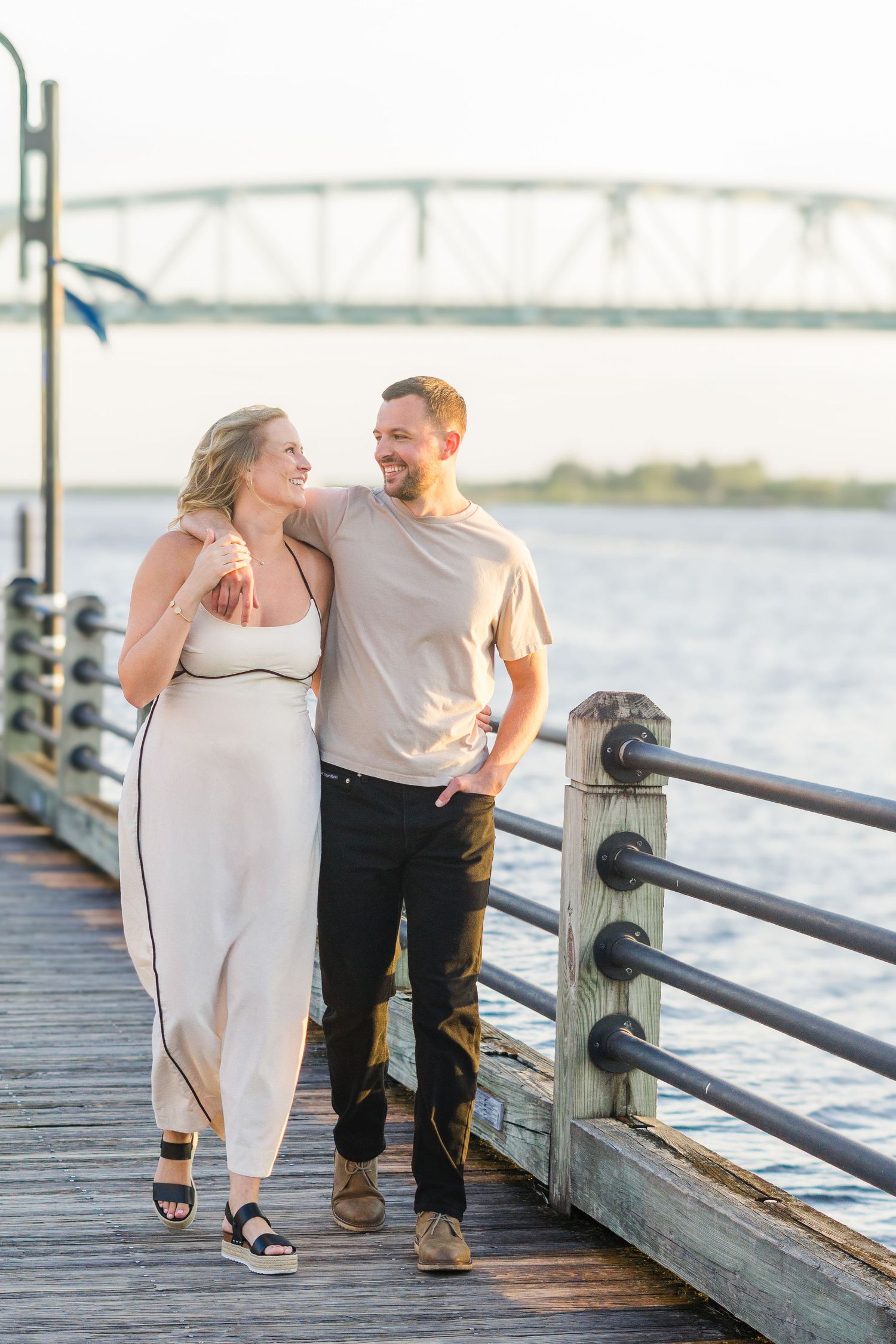 A couple walking and laughing together on a wooden pier with a bridge vista.