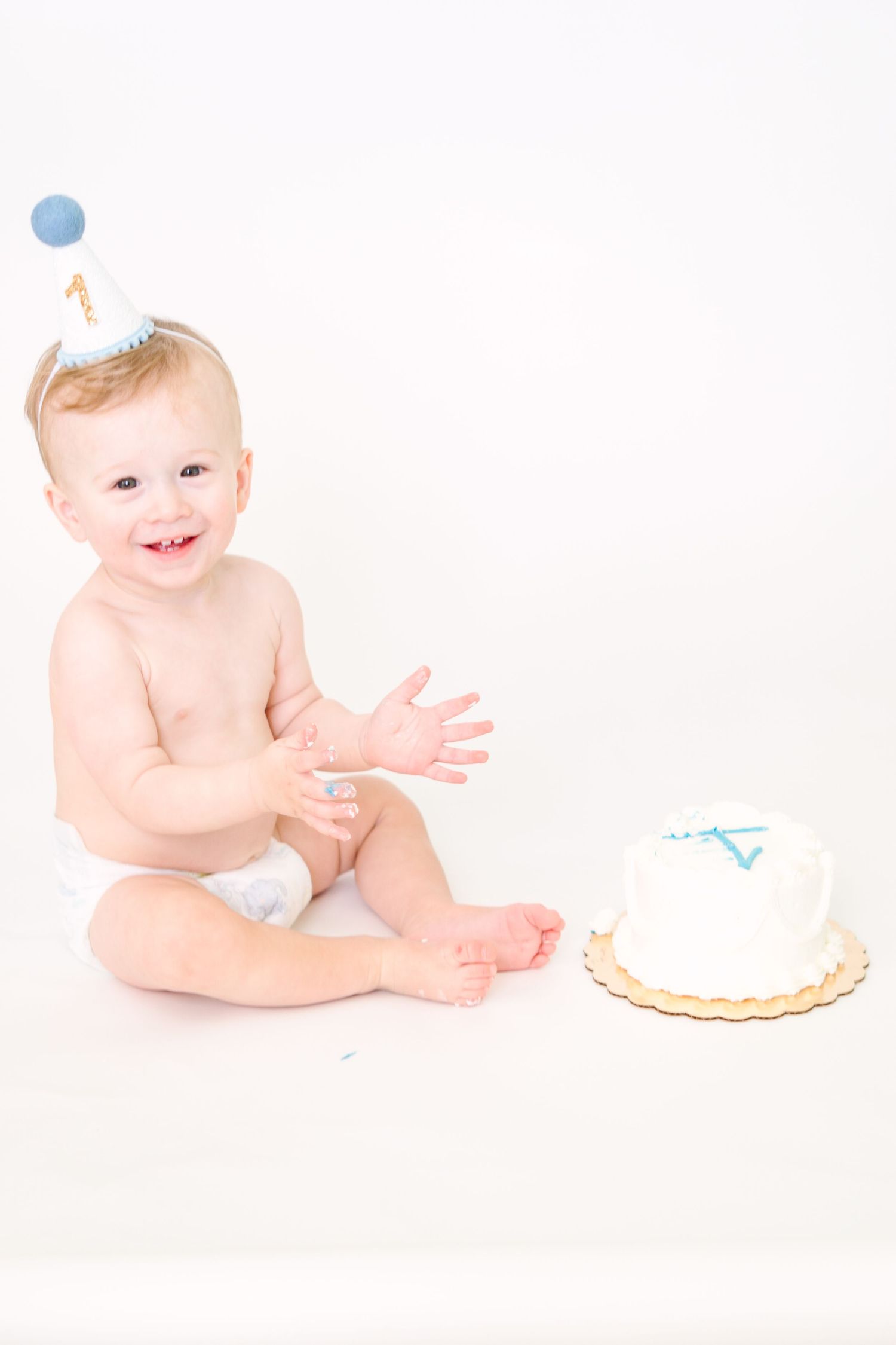 Baby in birthday hat sitting next to small white cake on white background.