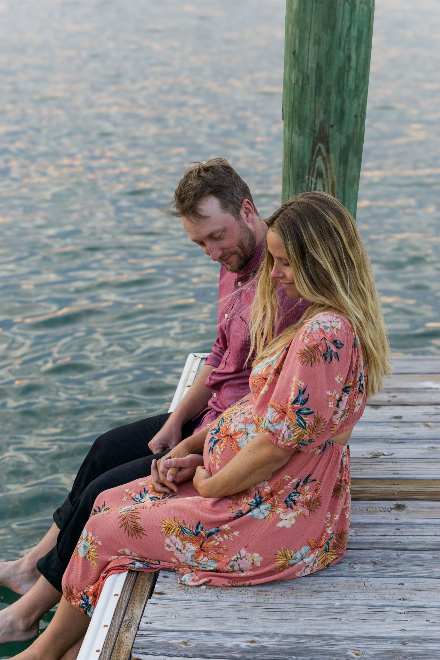 A couple gazes at each other while sitting on a wooden pier by the water.