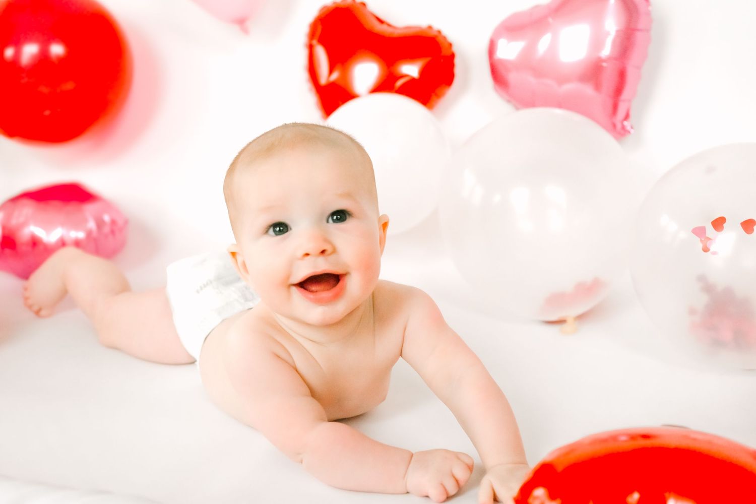 A baby surrounded by pink and red heart-shaped balloons in a light filled studio setting.