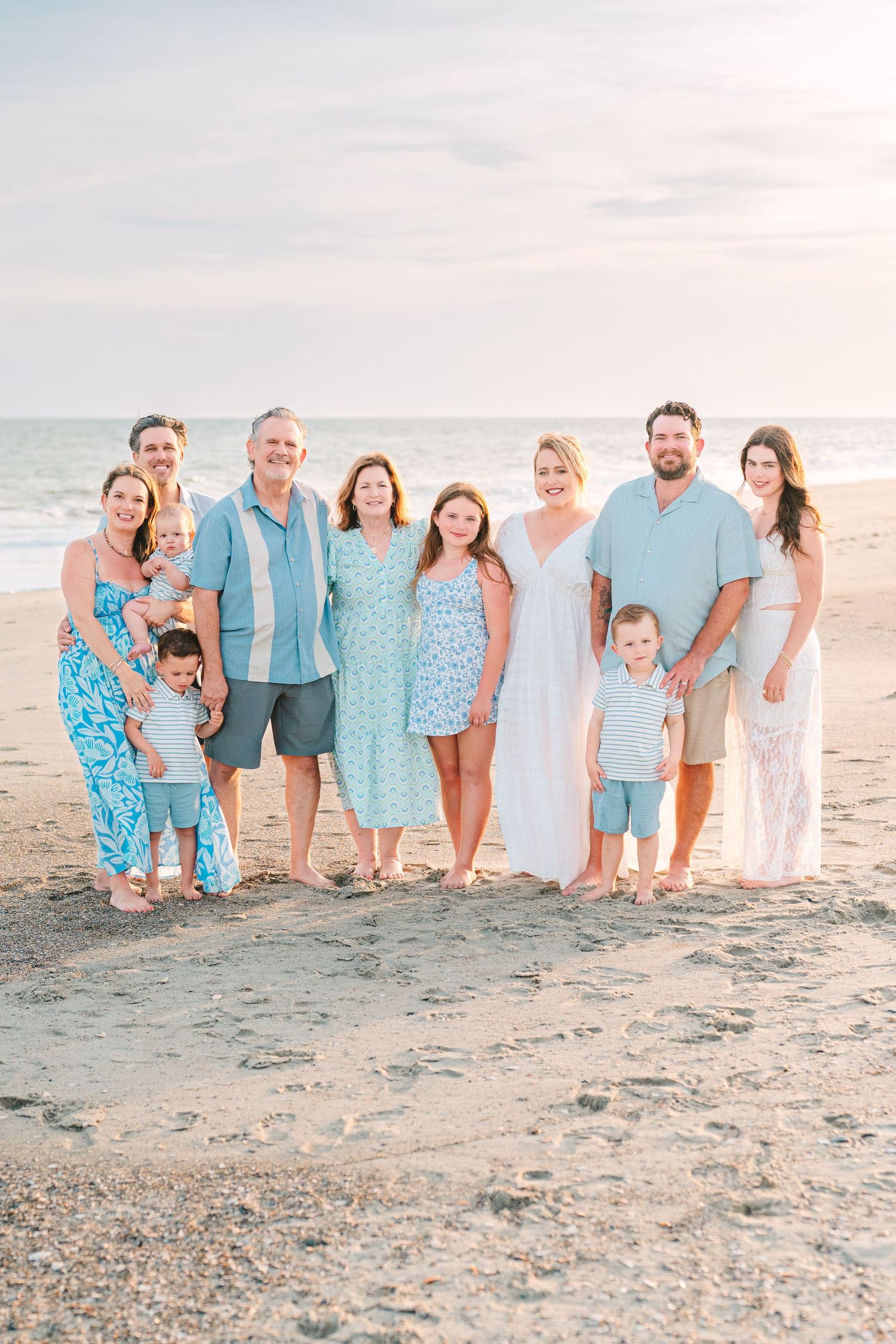 Extended family in coordinating blue and white outfits posing on beach at sunset.