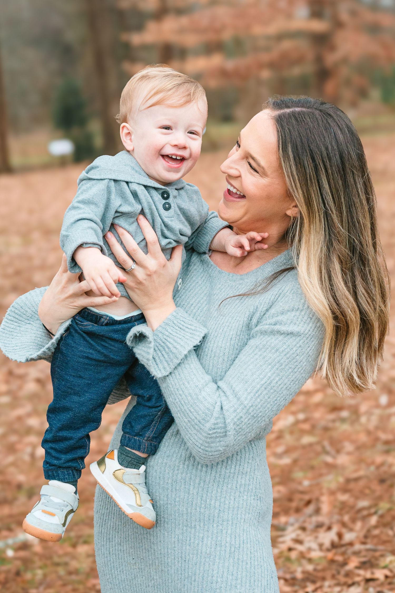 A parent in a gray sweater lifts and plays with a laughing toddler in a blue hoodie among fallen autumn leaves outdoors.