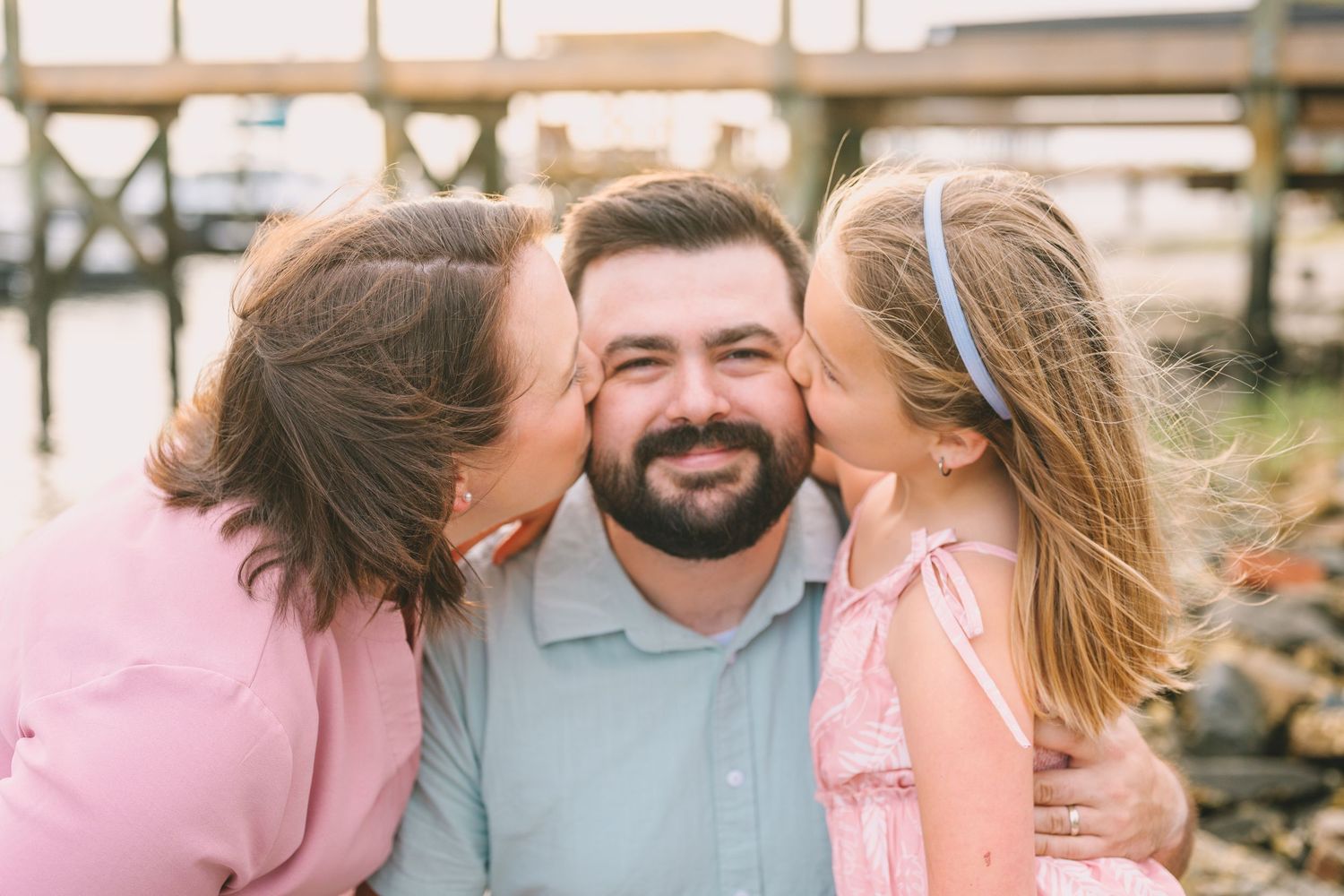 Sweet family moment captured outdoors wearing soft pink and gray tones with blurred outdoor background.