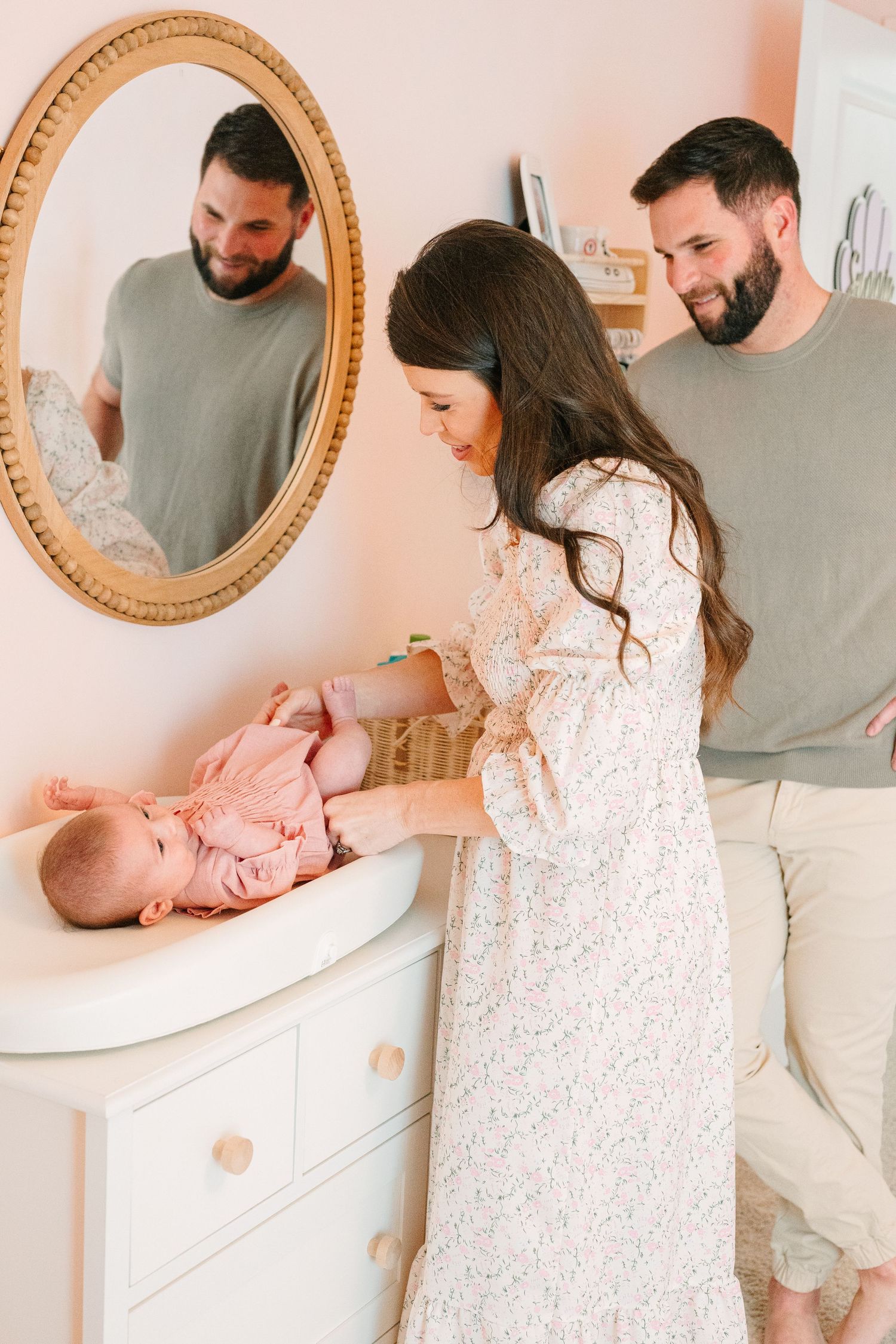 Baby lies on white changing table while being tended to in pink nursery room with ornate gold mirror.