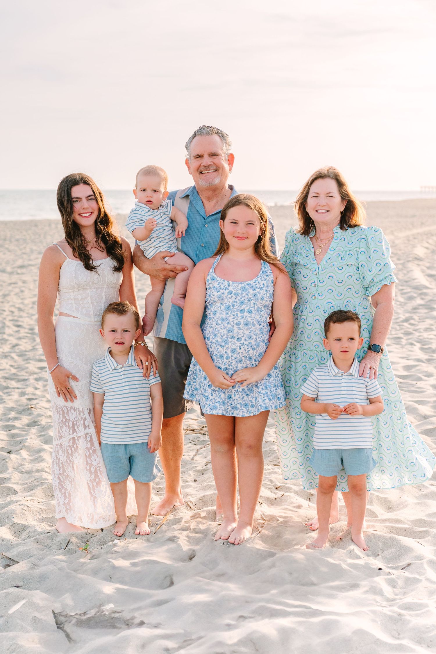 A family of six poses for a sunset beach portrait wearing coordinated blue and white coastal outfits.