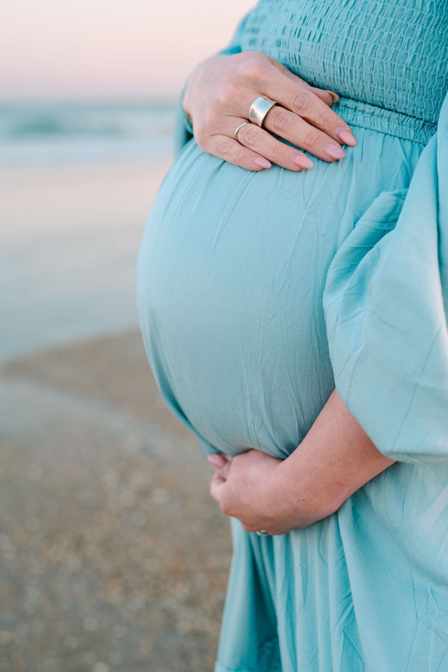 Close-up of pregnant woman in turquoise dress cradling her baby bump on a beach at sunset, wearing wedding ring.