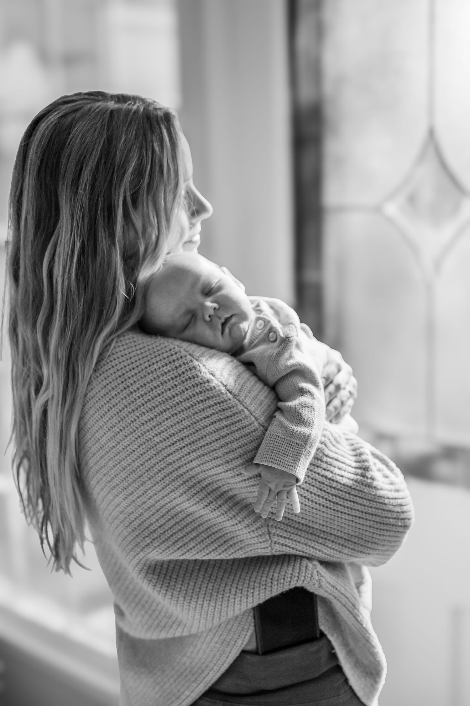 A tender black and white portrait of a parent holding a sleeping baby against their shoulder near a window.