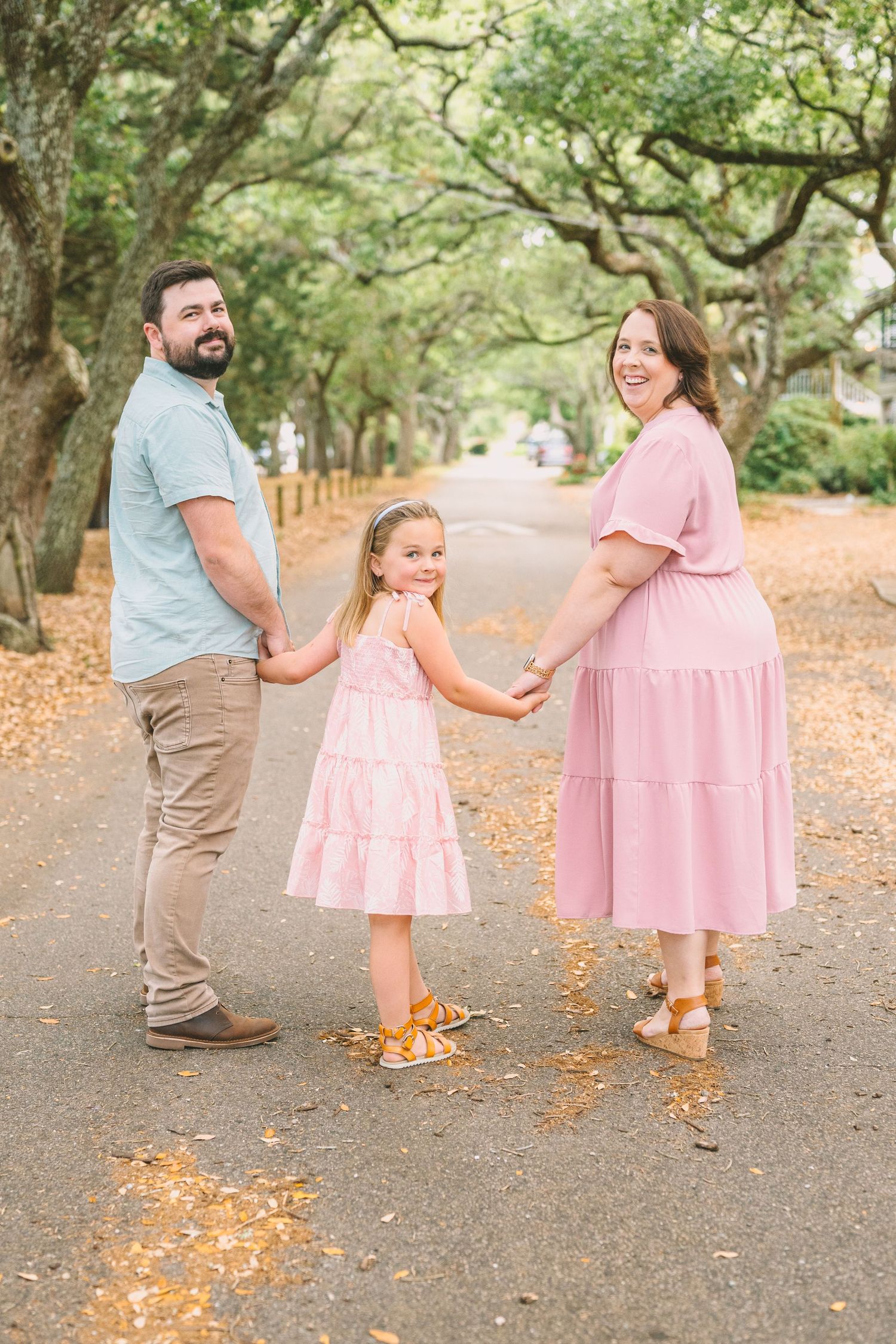 A family walks together on a tree-lined path, with mother and daughter wearing matching pink dresses.