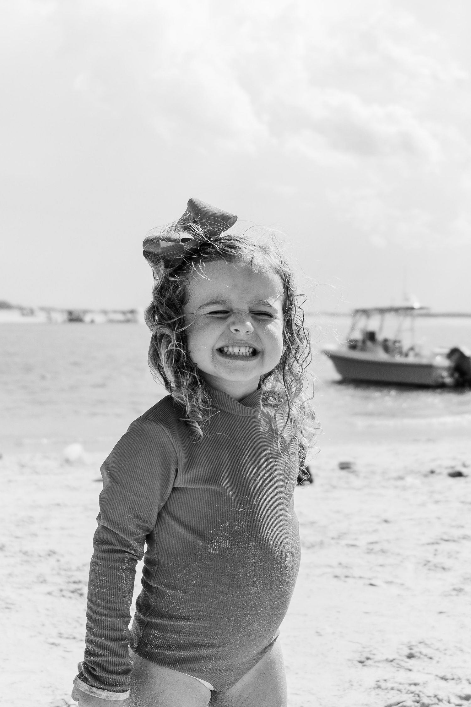 Candid black and white portrait of child with curly hair smiling on beach with boat in background.