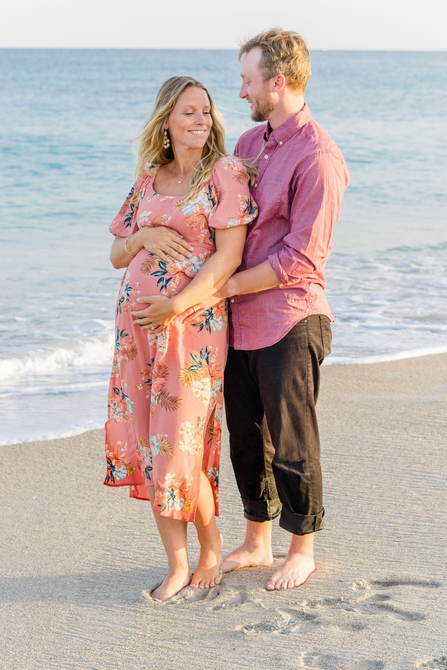 Happy couple embraces on beach during sunset maternity session in coordinating pink and coral outfits.