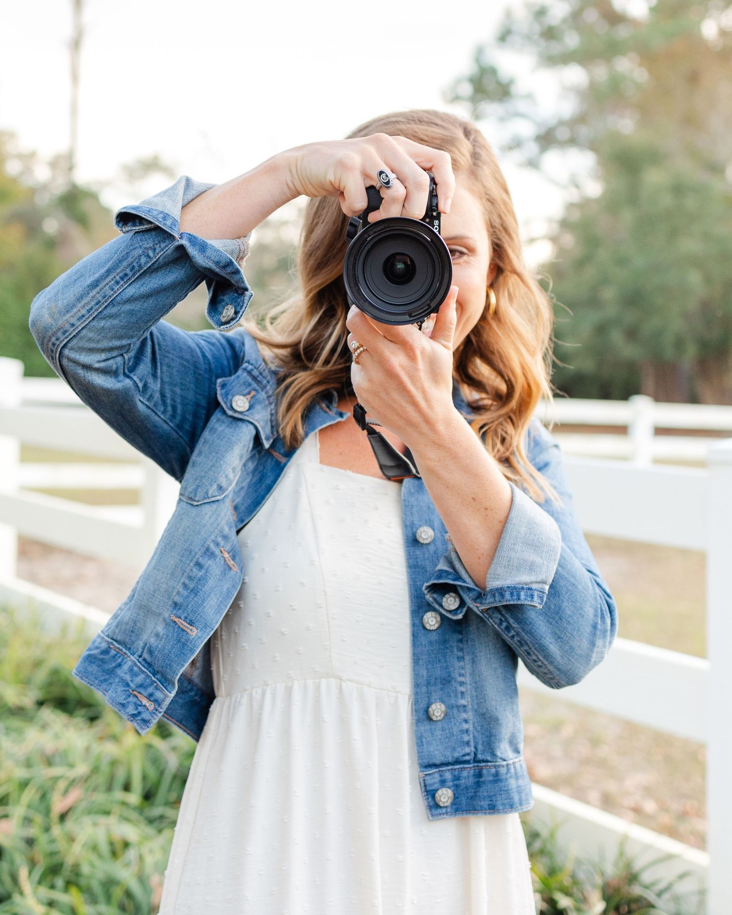Photographer in white dress and jean jacket taking pictures with professional camera near white fence.