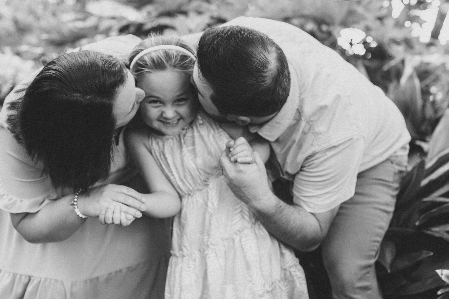 A sweet black and white photo captures an intimate family moment with parents embracing a giggling child outdoors.