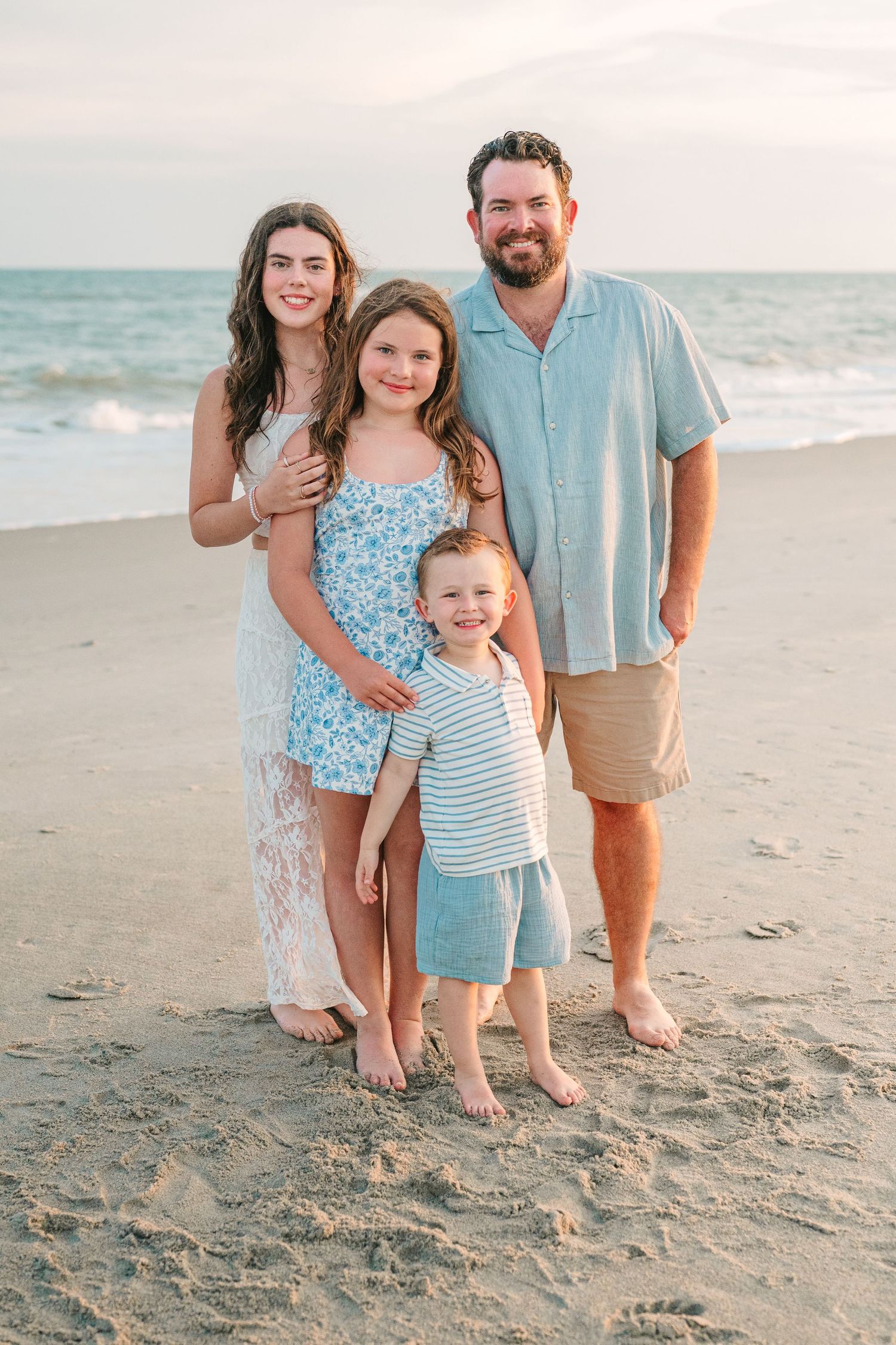 Family of four in coordinating blue and white outfits pose on beach at sunset.