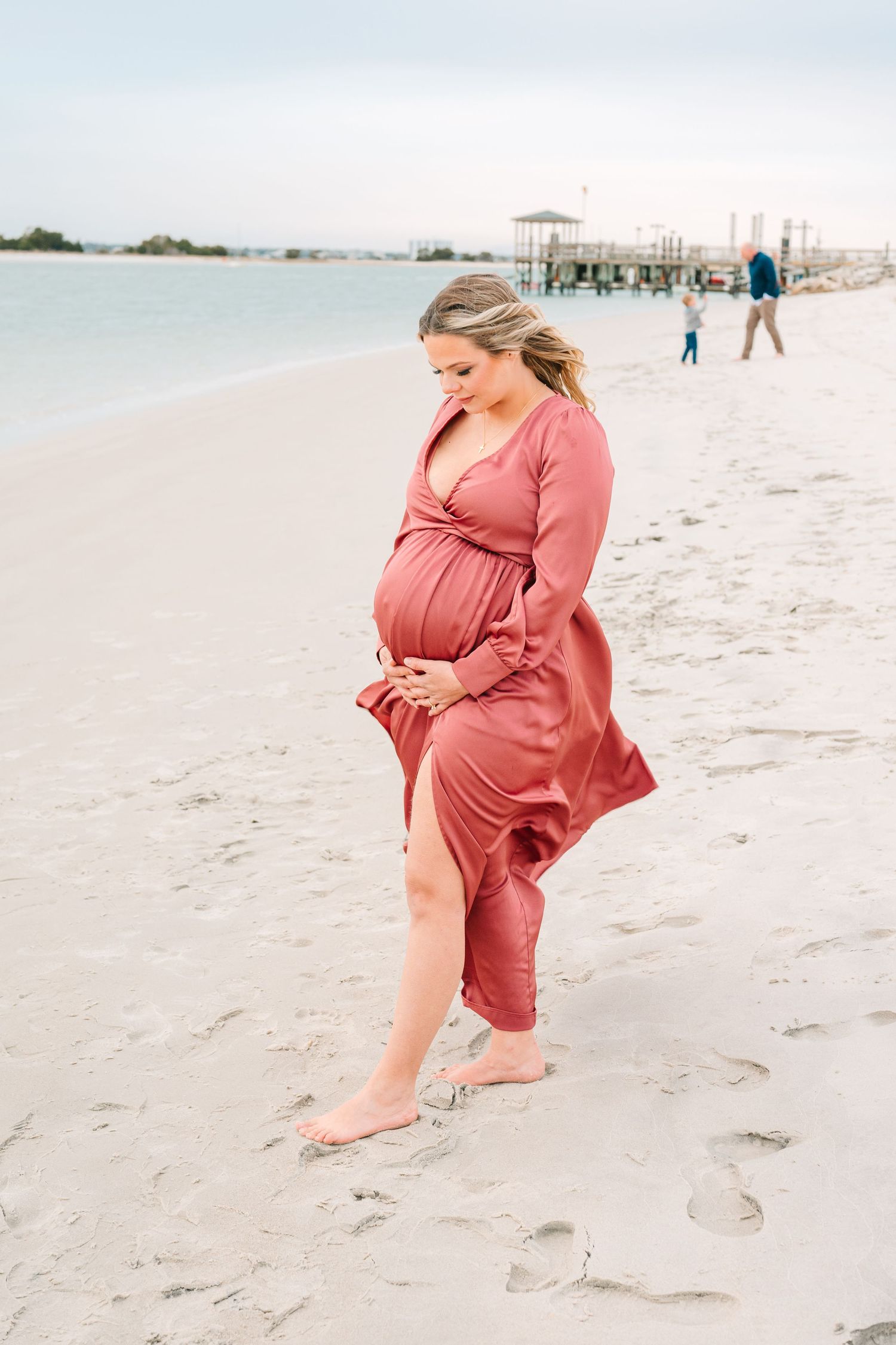 Pregnant woman in flowing coral dress walks barefoot along white sandy beach with pier in background during sunset.