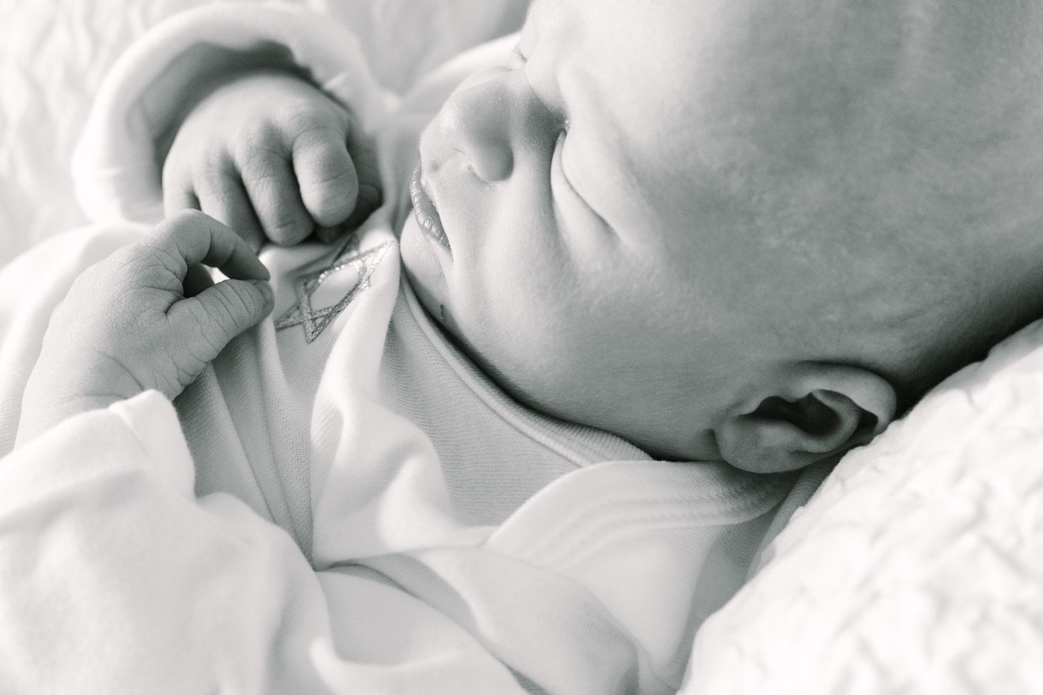Close-up black and white photograph of a sleeping baby swaddled in soft white fabric, showing tiny hand and ear details.