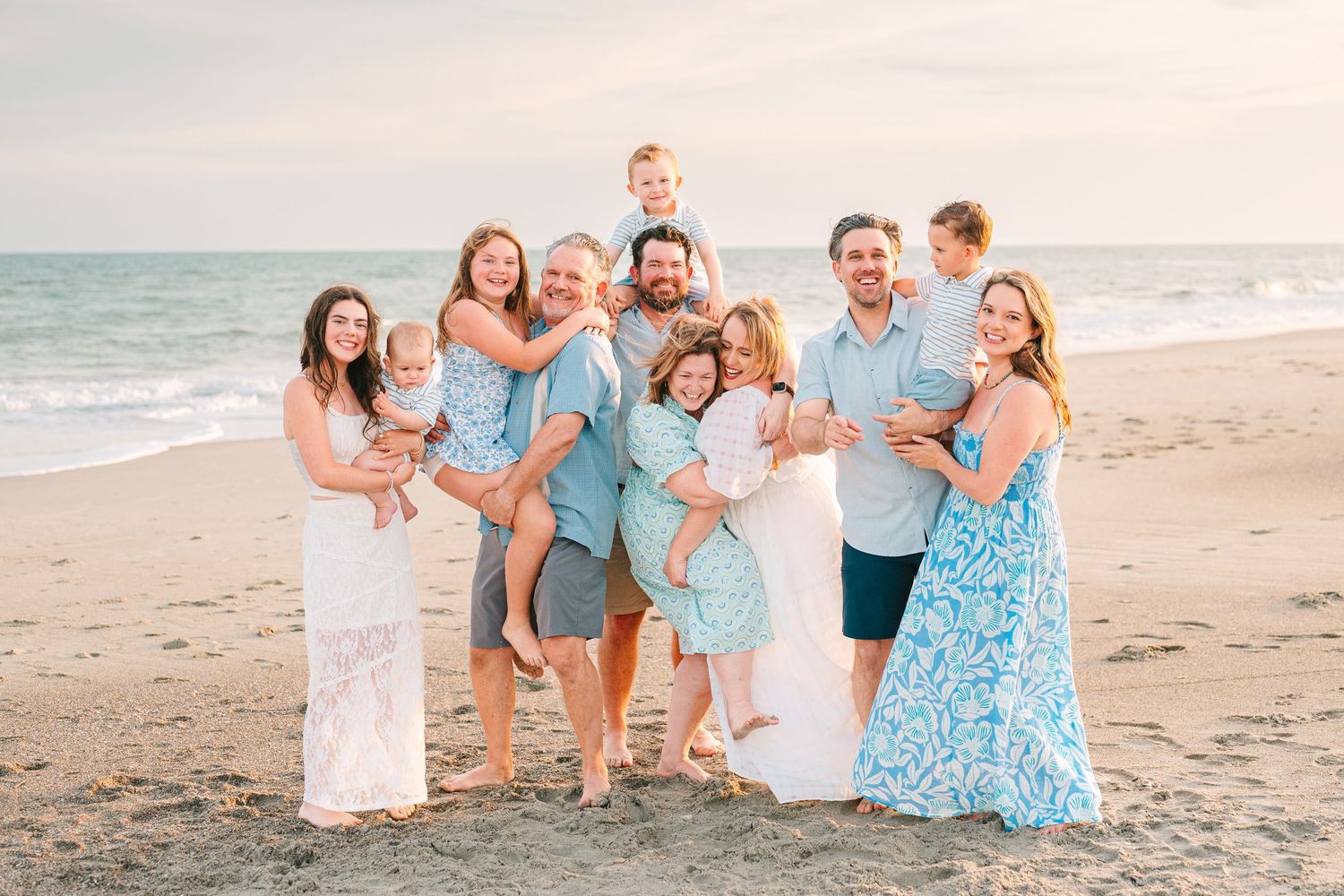 Large family group shares joyful moment together on beach wearing coordinated blue and white outfits.