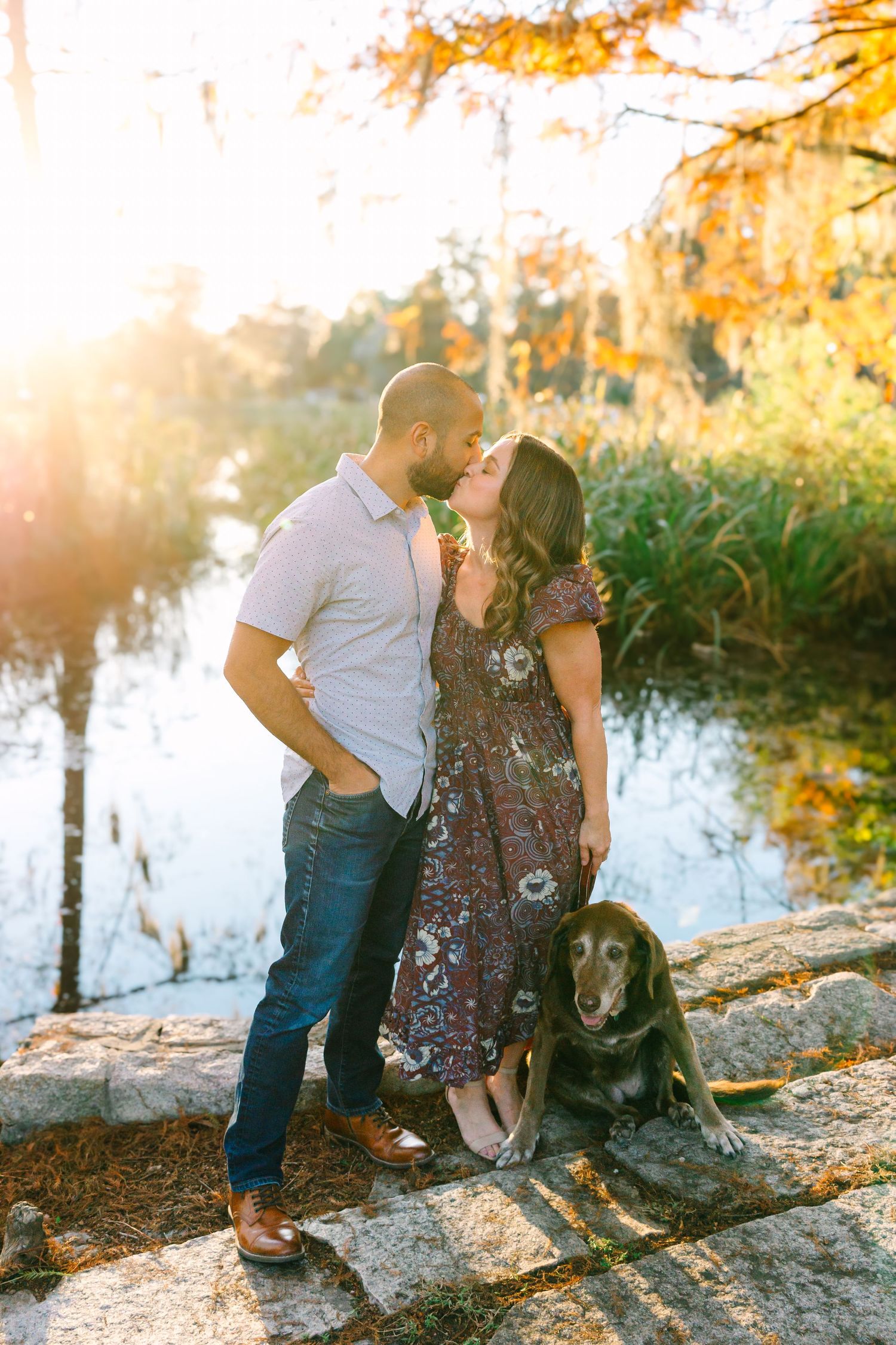 Romantic couple with dog by the water during golden sunset among autumn trees.