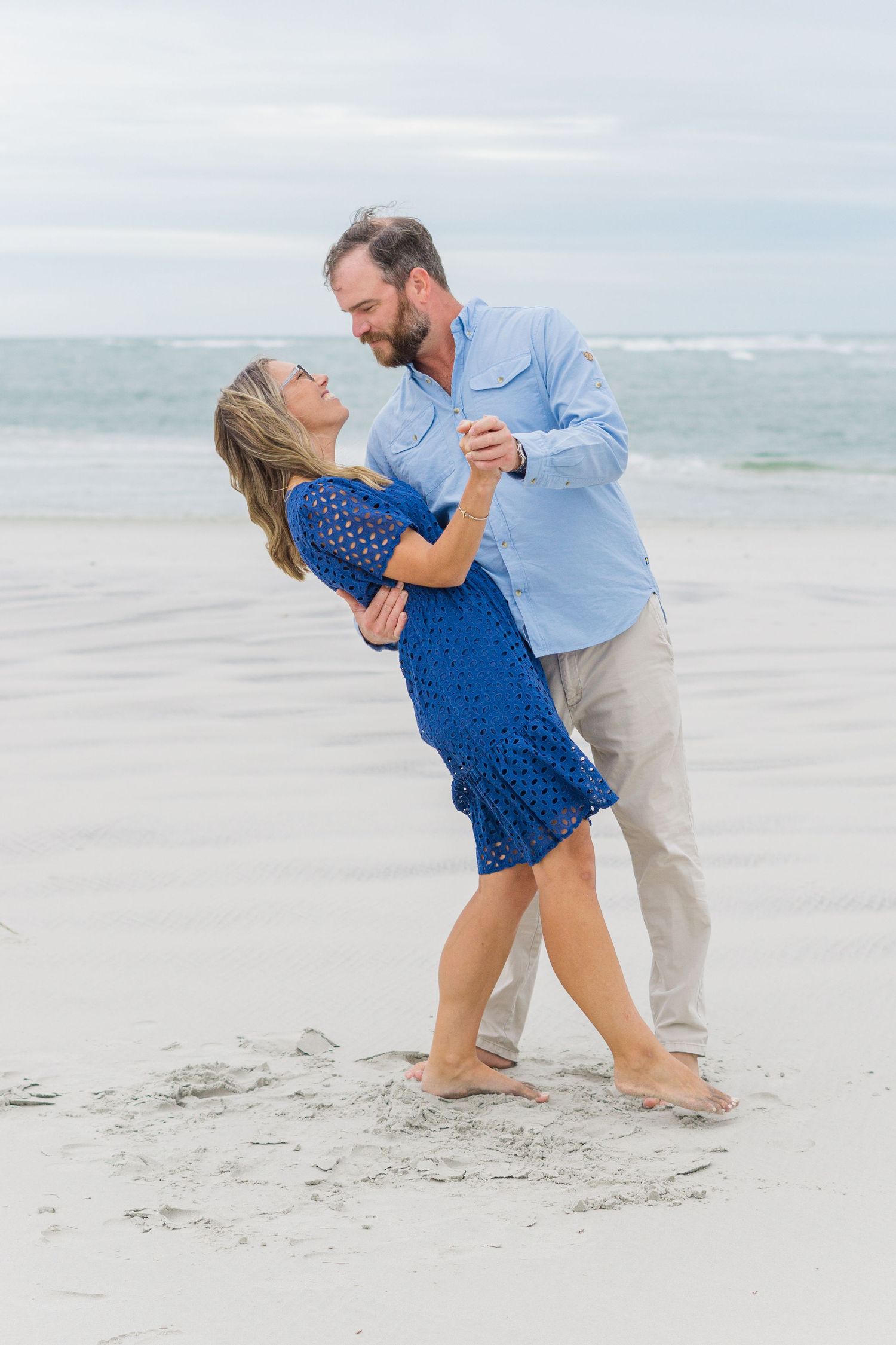 A couple dances playfully on the beach with ocean waves in the background.