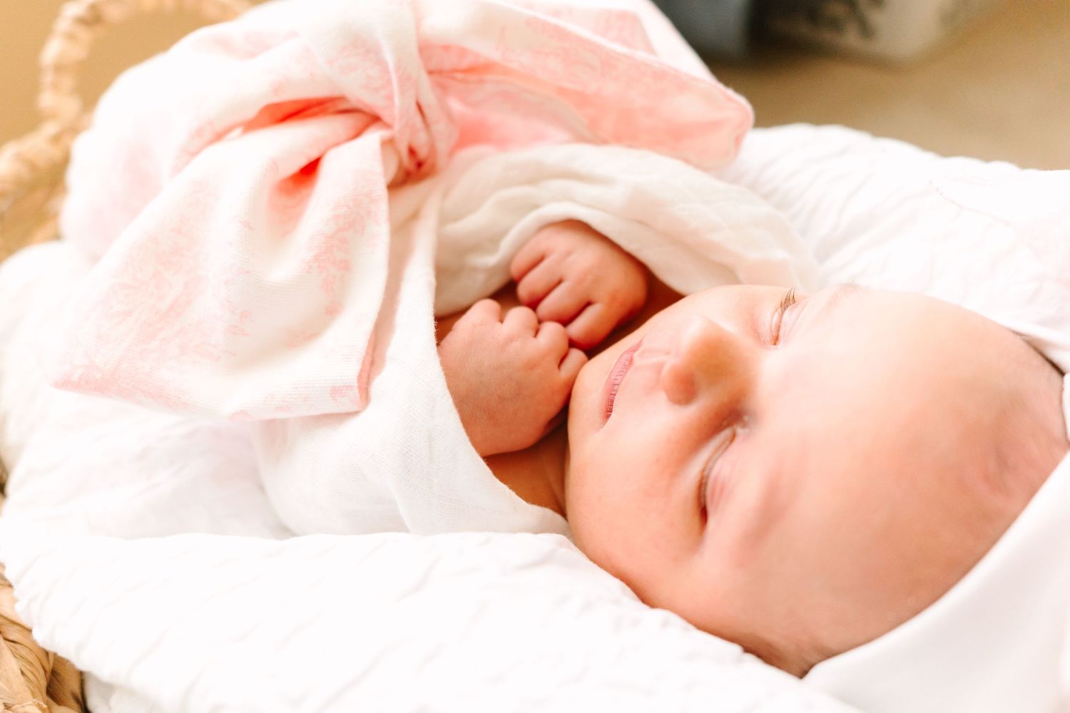 A sleeping newborn with pink bow accessory resting in a woven basket with soft lighting.