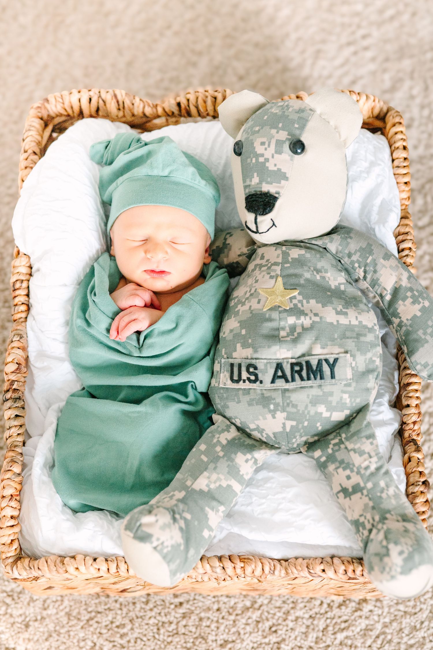 Newborn in mint outfit lies beside plush Army teddy bear in military uniform in woven basket.