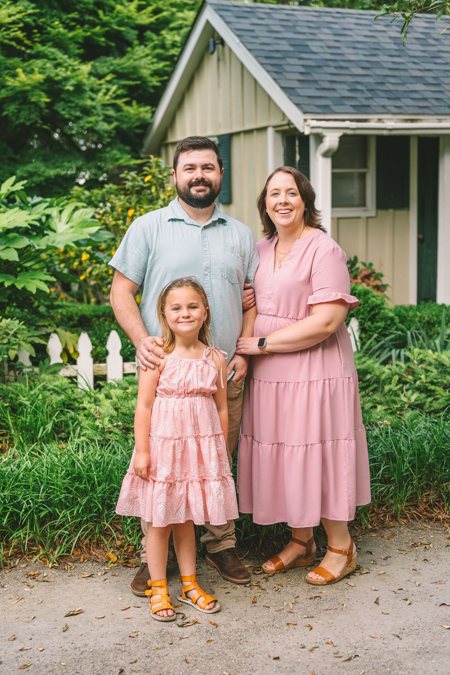 A family stands together outdoors in coordinated pink outfits in front of a rustic cottage with garden landscaping.