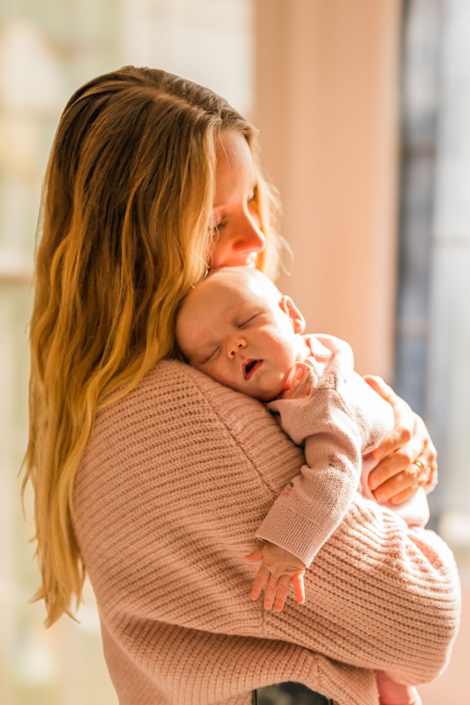 A tender moment as a mother in a pink sweater cuddles her sleeping baby in warm sunlit room.