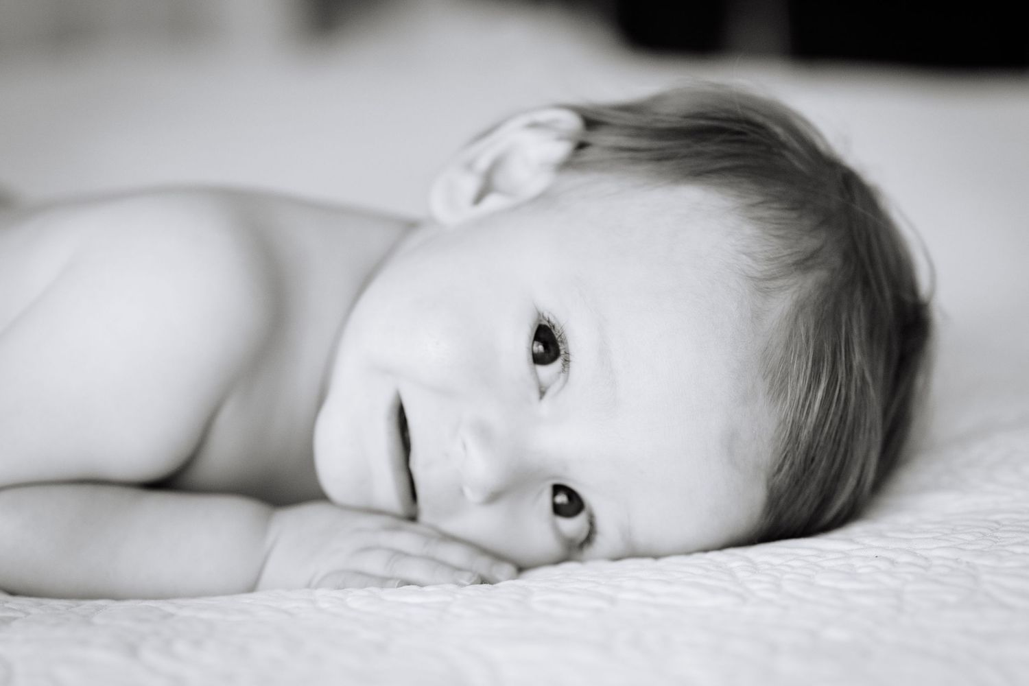 A black and white portrait of a young child lying on white bedding with soft lighting.