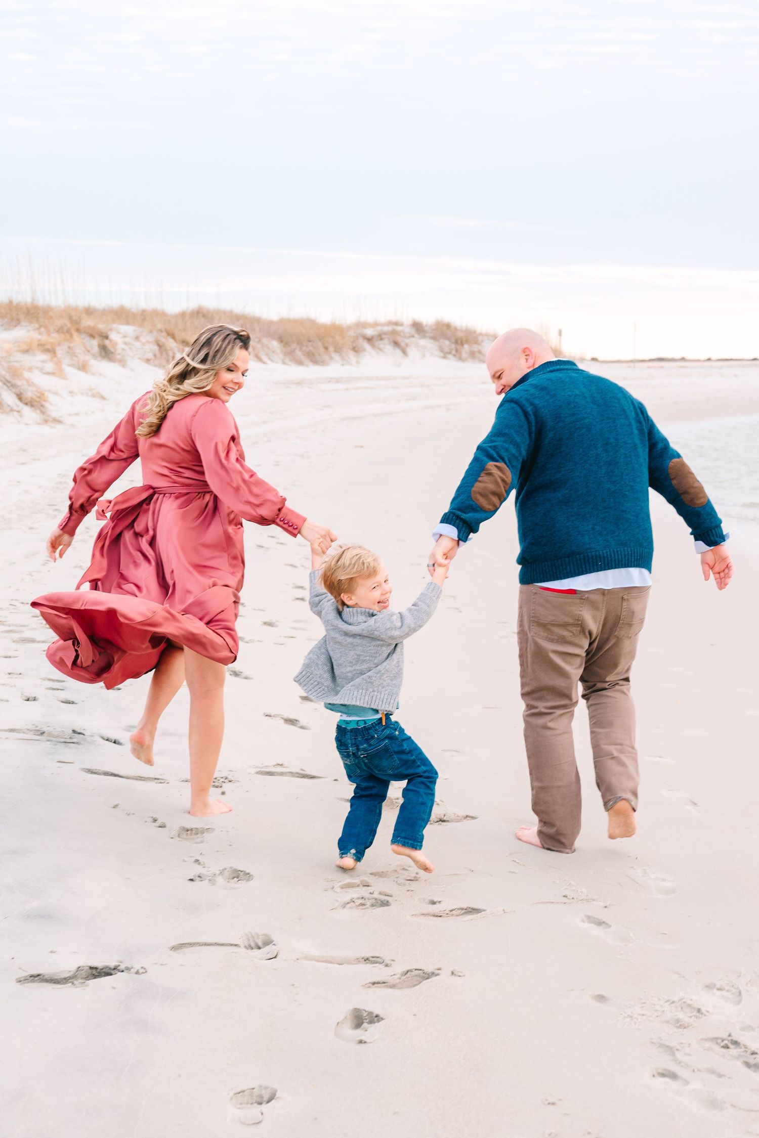 A family holds hands while walking barefoot together on a sandy beach during a breezy day.