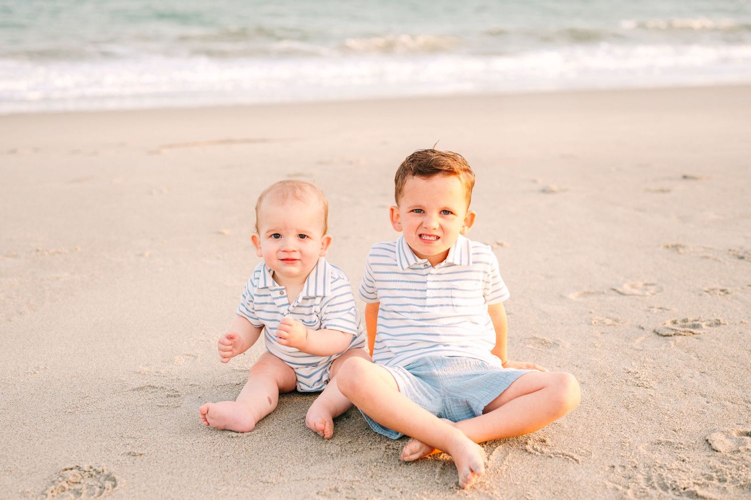 Two young children in matching striped outfits sit together on sandy beach.