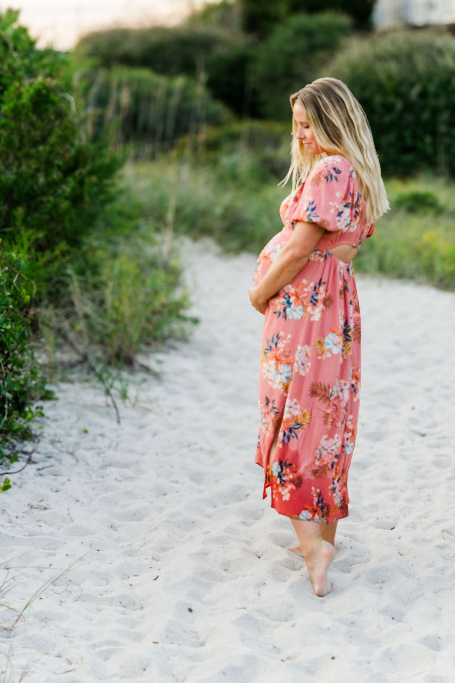Expecting mother in coral floral dress standing barefoot on a sandy beach path.