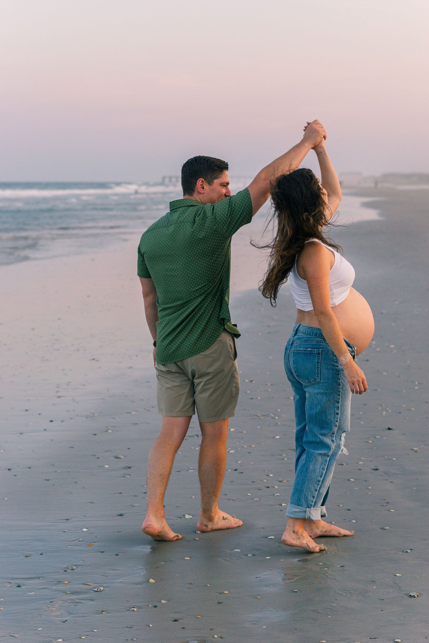 A couple dances together on the beach during golden hour, celebrating their upcoming arrival.