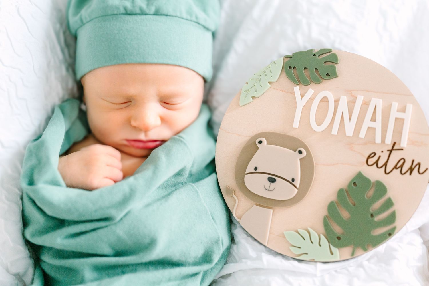Sleeping newborn in mint green wrap next to round wooden name sign with safari animal design.