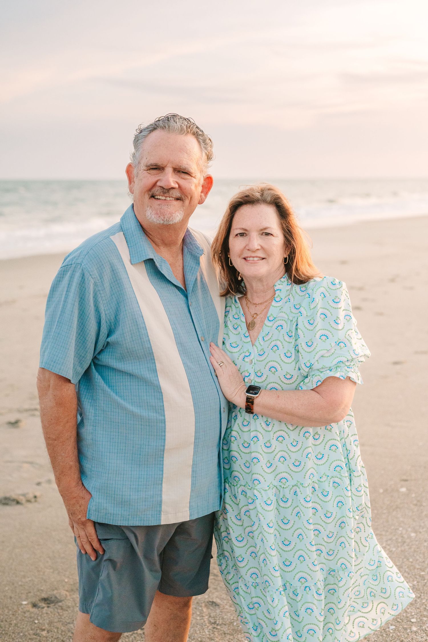 A couple in coordinating blue outfits poses together on the beach at sunset.