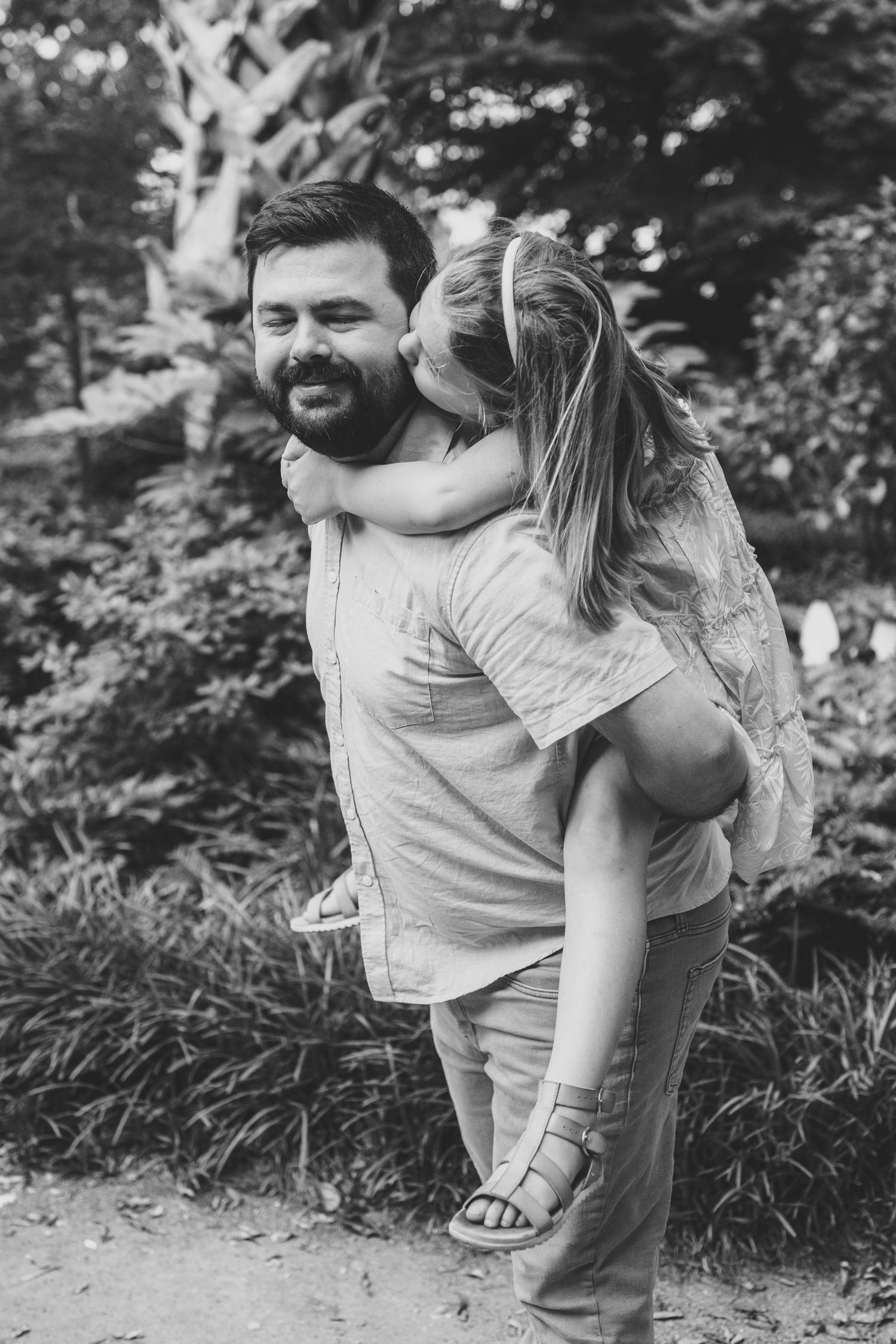 A person gives a piggyback ride to a child while walking outdoors on a nature path in black and white photography.