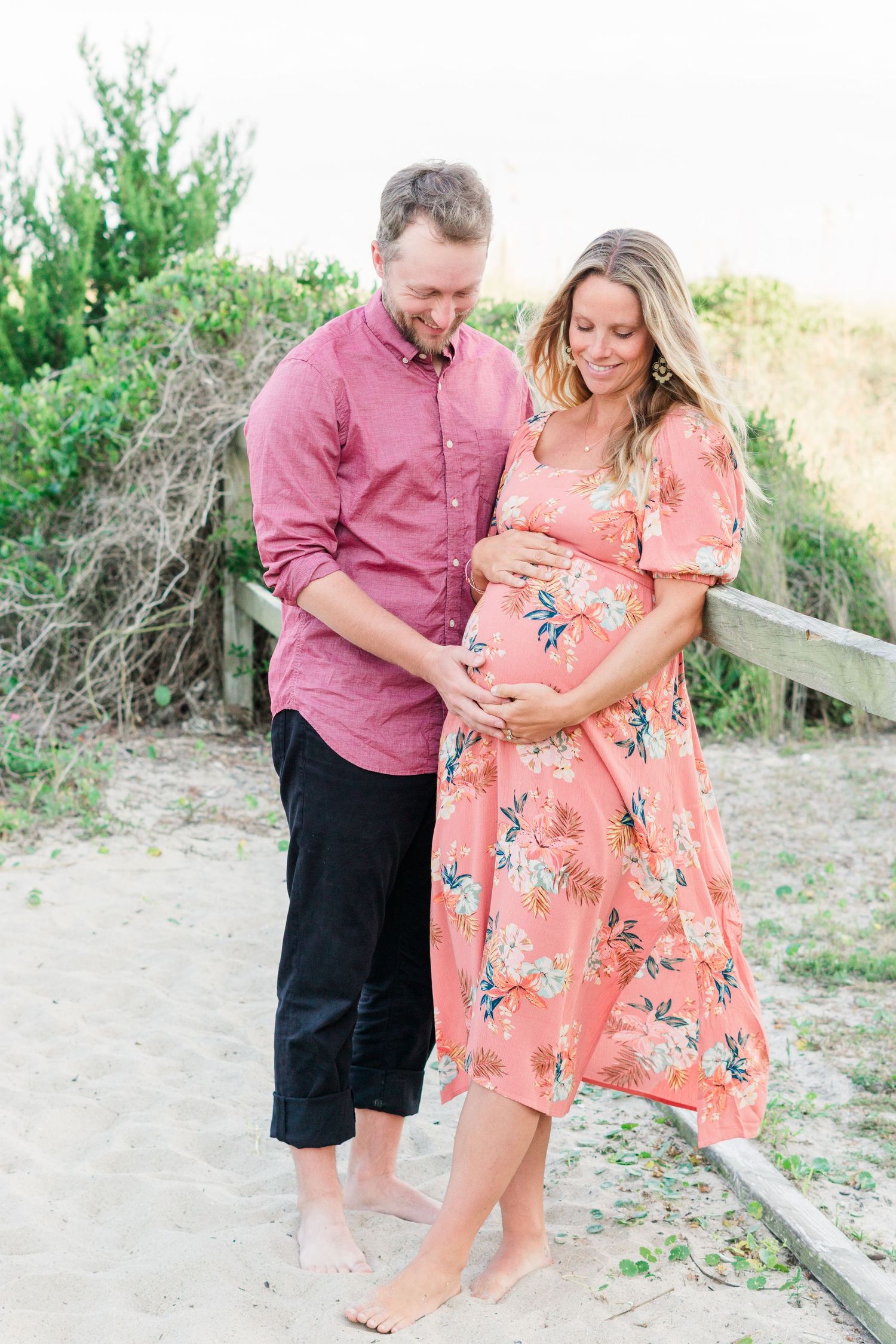 Maternity photo of couple in coordinated outfits on sandy beach path.
