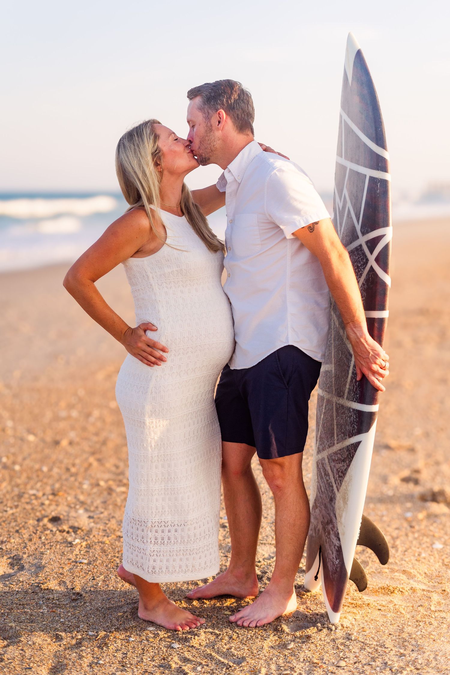 Expecting couple shares intimate moment by surfboard during beach sunset session.