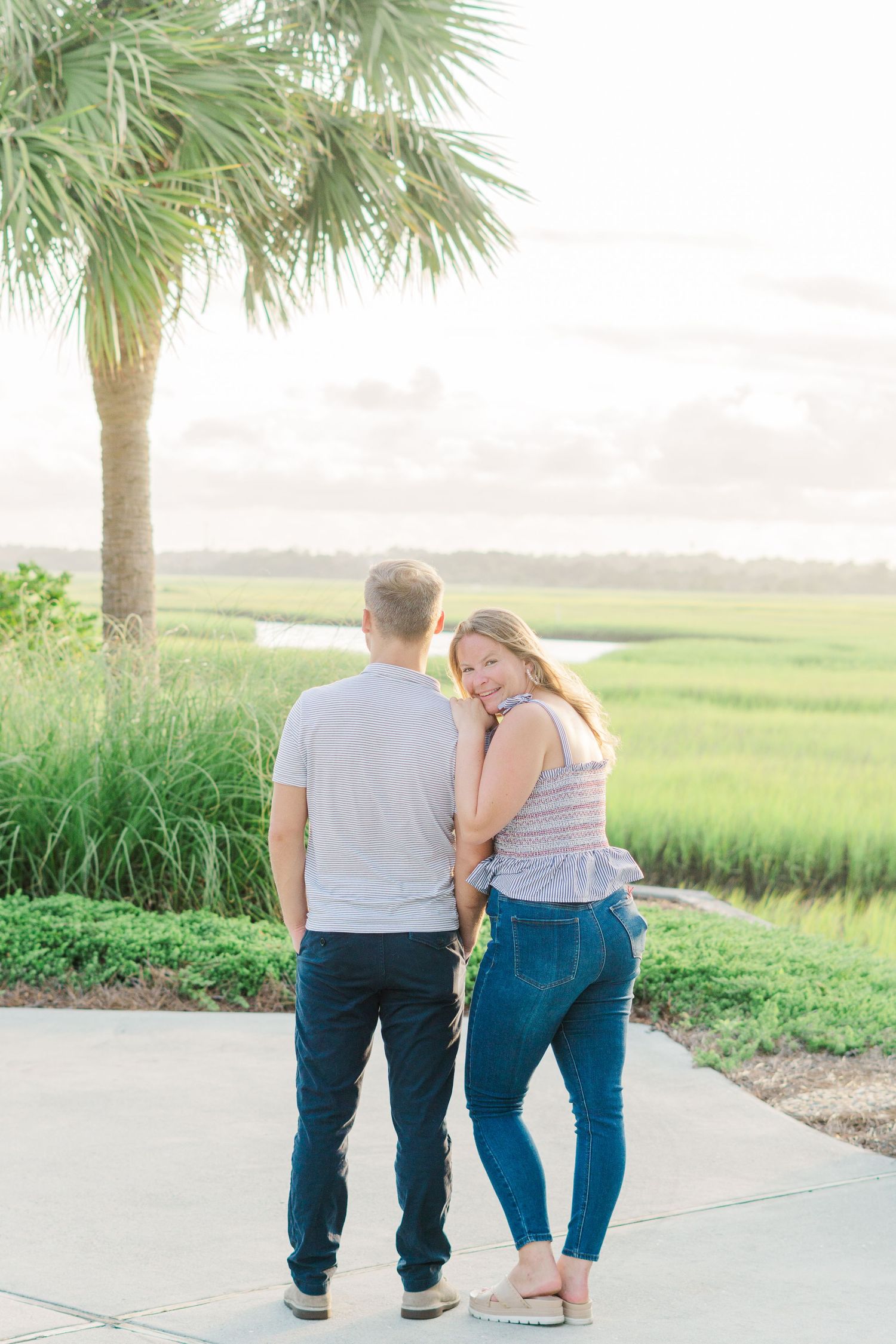 Couple shares tender moment under palm tree overlooking coastal marsh landscape.