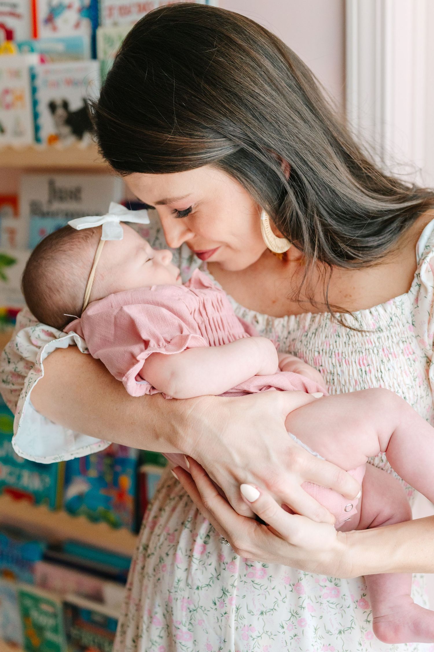 Mother in floral dress tenderly holding sleeping newborn baby wearing pink romper near bookshelf.