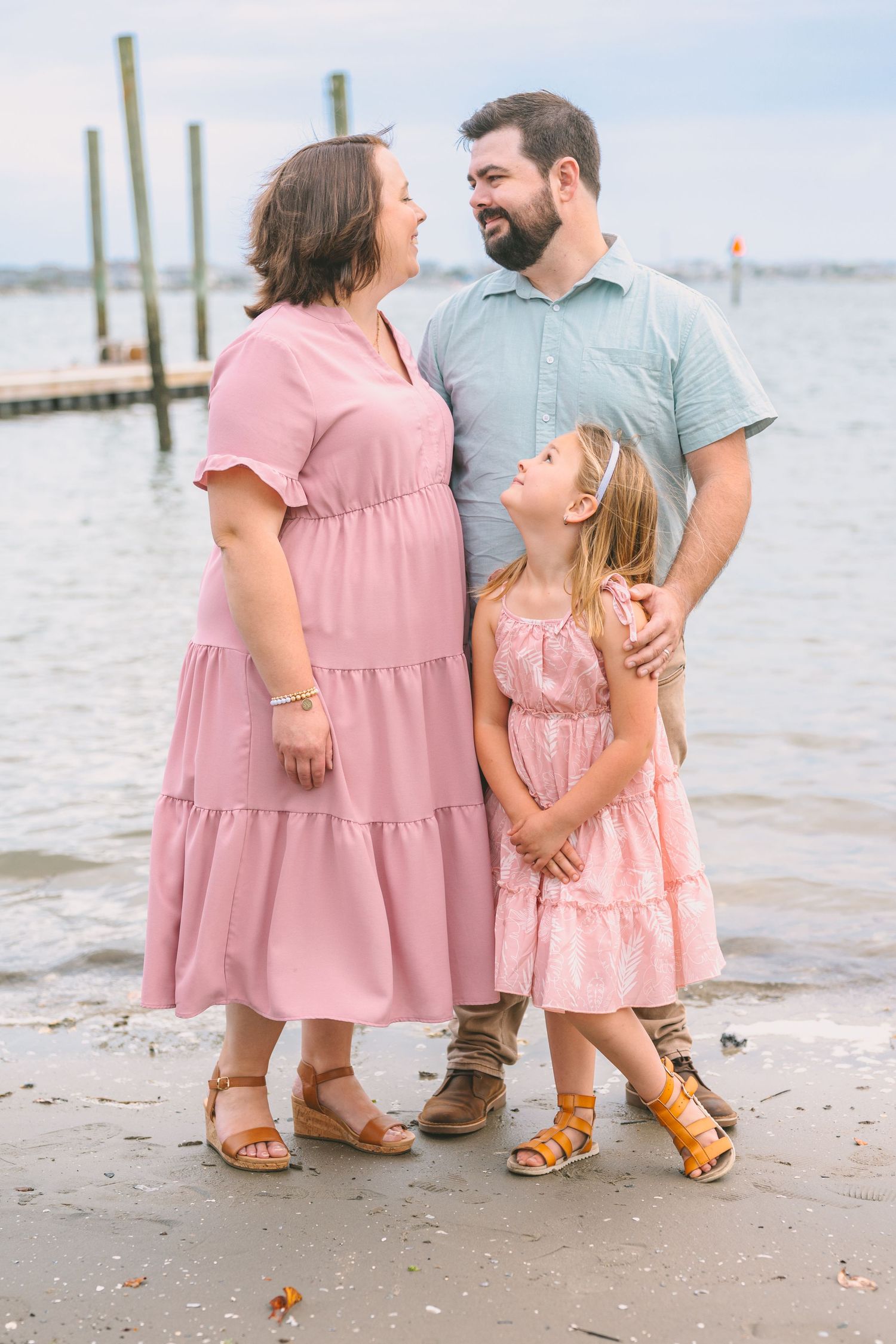 A family in matching pink outfits shares a tender moment together on a beach dock with water views.