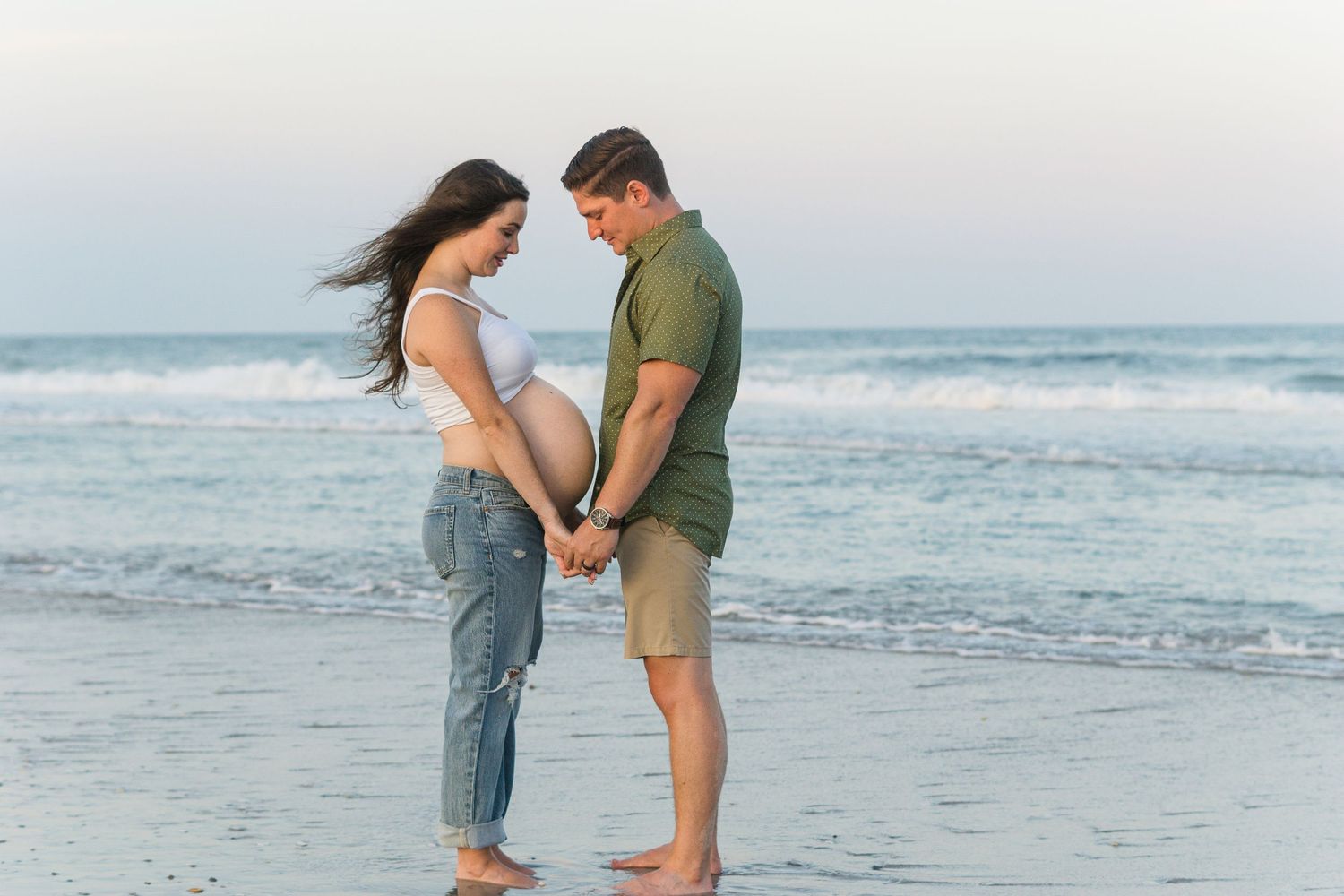 Beach maternity photo showing couple holding hands by ocean waves.