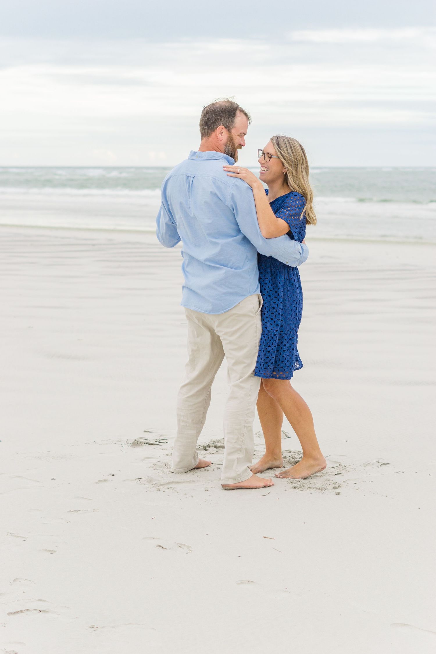 A couple in coordinating blue and khaki outfits dances barefoot on a pristine beach.