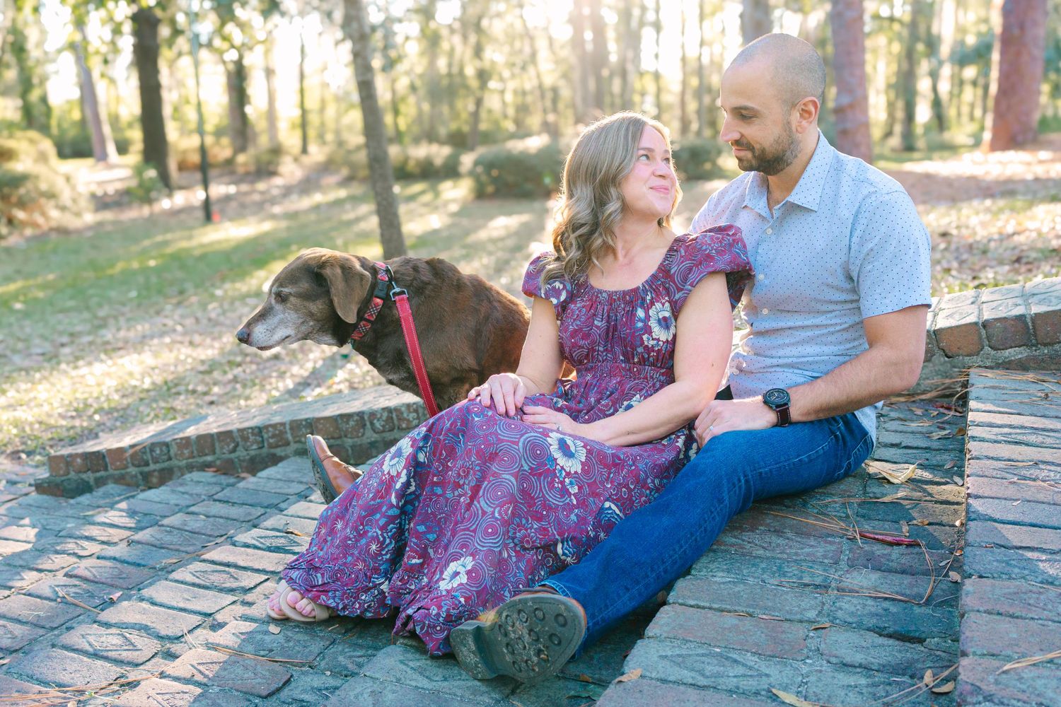 Couple sitting with their dog on brick pavers in a sunlit wooded area.