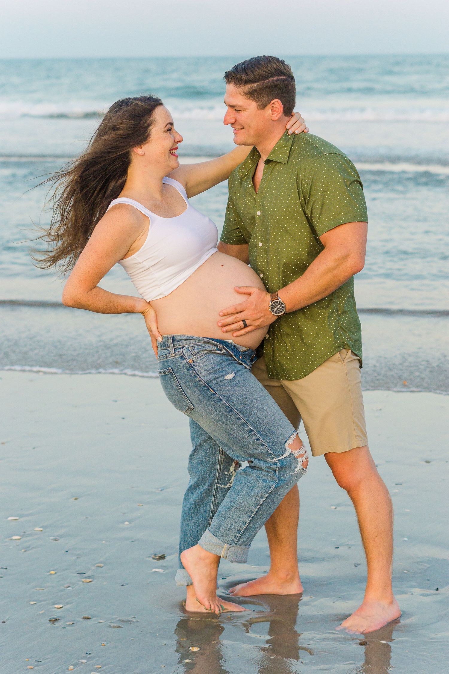 A joyful maternity photo shows a couple laughing together by the ocean at sunset.