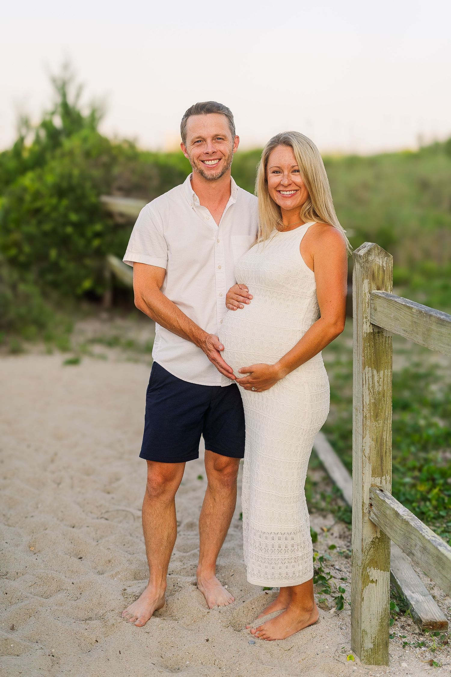 Expecting couple poses barefoot on beach path in matching casual white outfits.