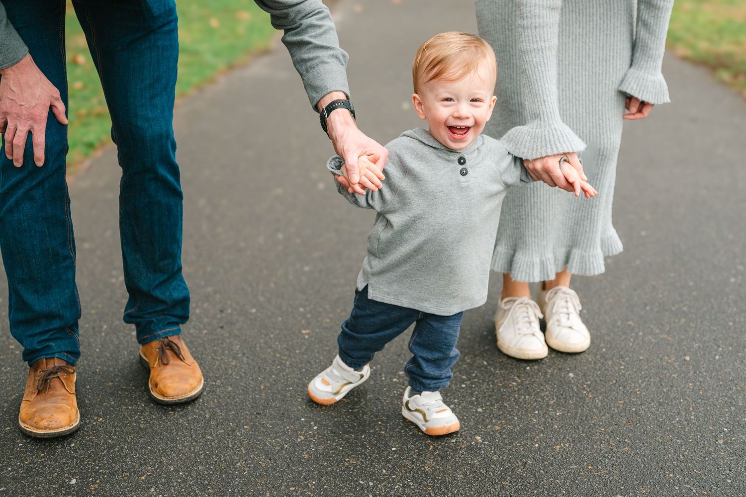 A toddler in a gray sweater taking steps while holding hands with adults on a paved pathway.