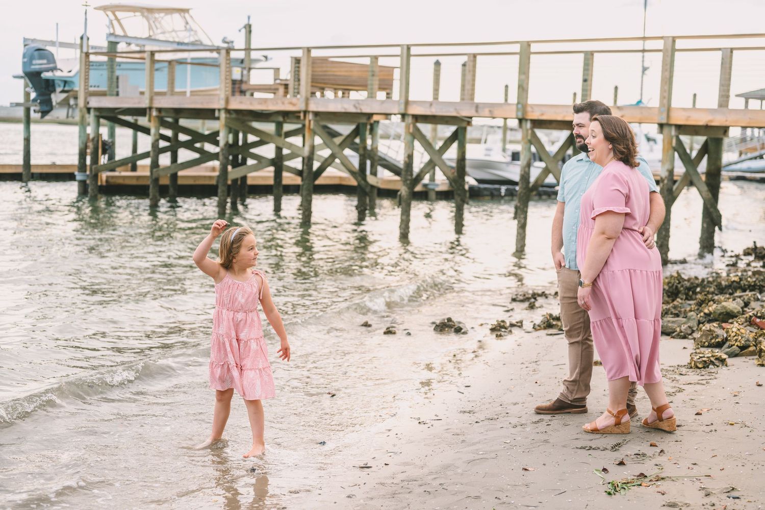Family in pink dresses enjoys a day at the waterfront with wooden pier and rocky shoreline in the background.