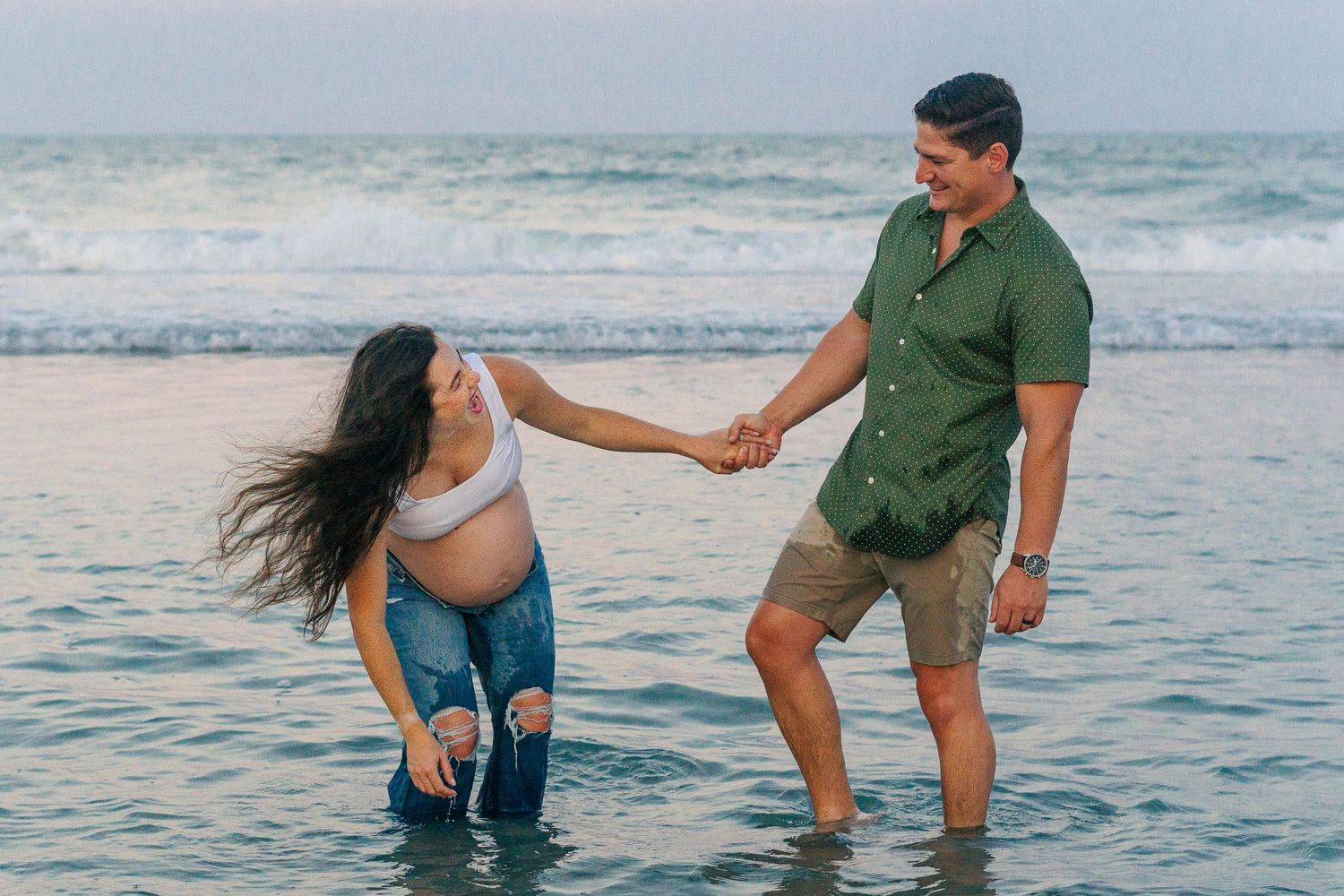 Playful couple holding hands while dancing in shallow ocean waters at dusk.