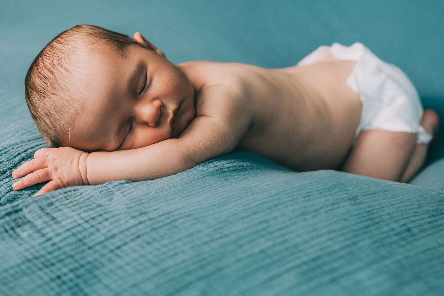 Sleeping infant wearing white diaper rests peacefully on teal-colored textured blanket.