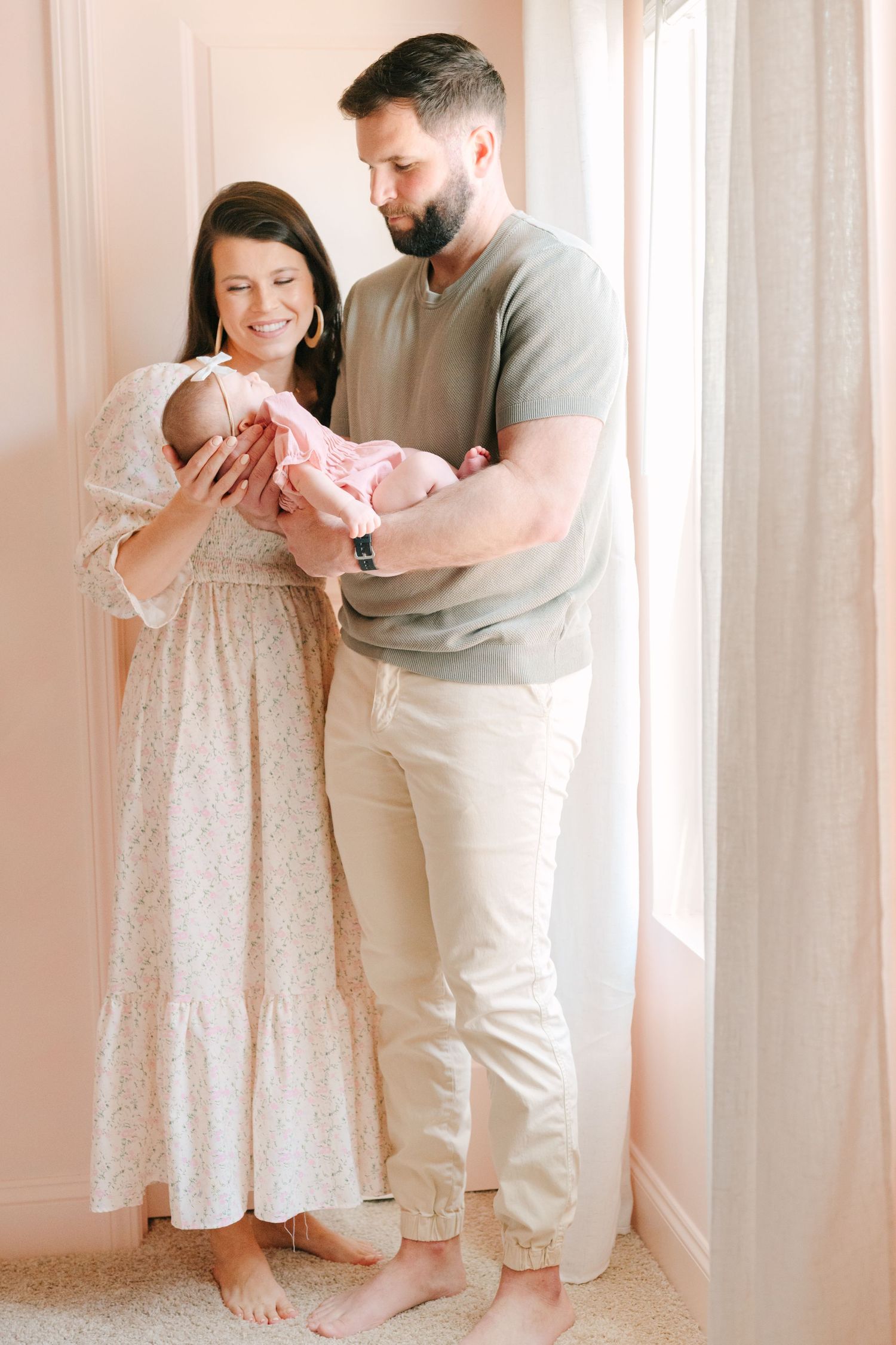 A couple in casual attire lovingly holds their newborn baby next to a sunlit window in a peaceful home setting.
